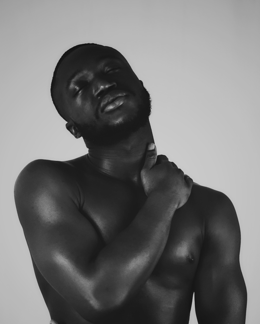 Black and white portrait of a shirtless person with eyes closed, head tilted back, hand on neck, neutral background.