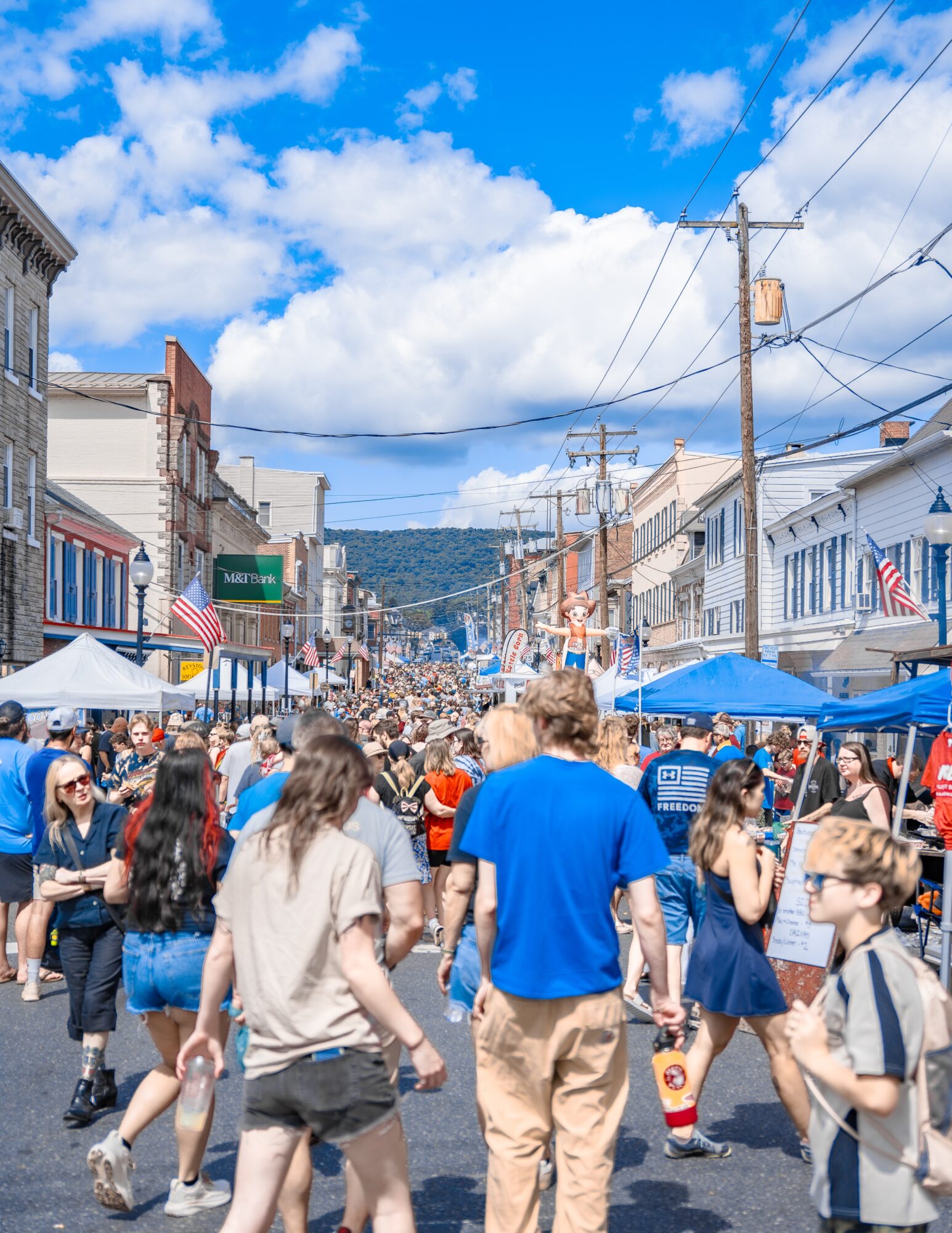 Crowded street with people walking, shops, tents, and blue sky with clouds, in a small town or city.