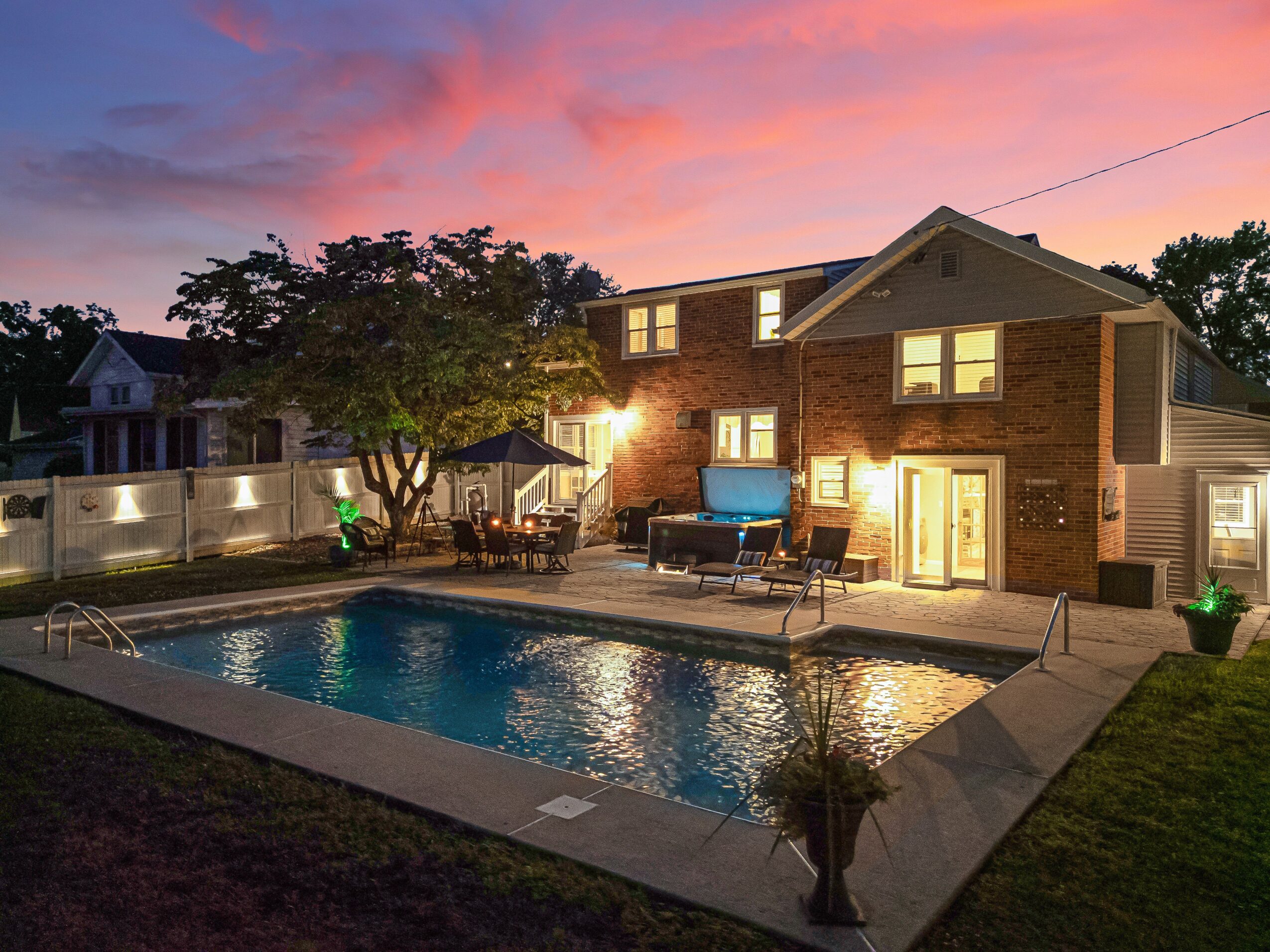 Backyard with swimming pool, patio furniture, house with lit windows, trees, and a colorful sunset sky.