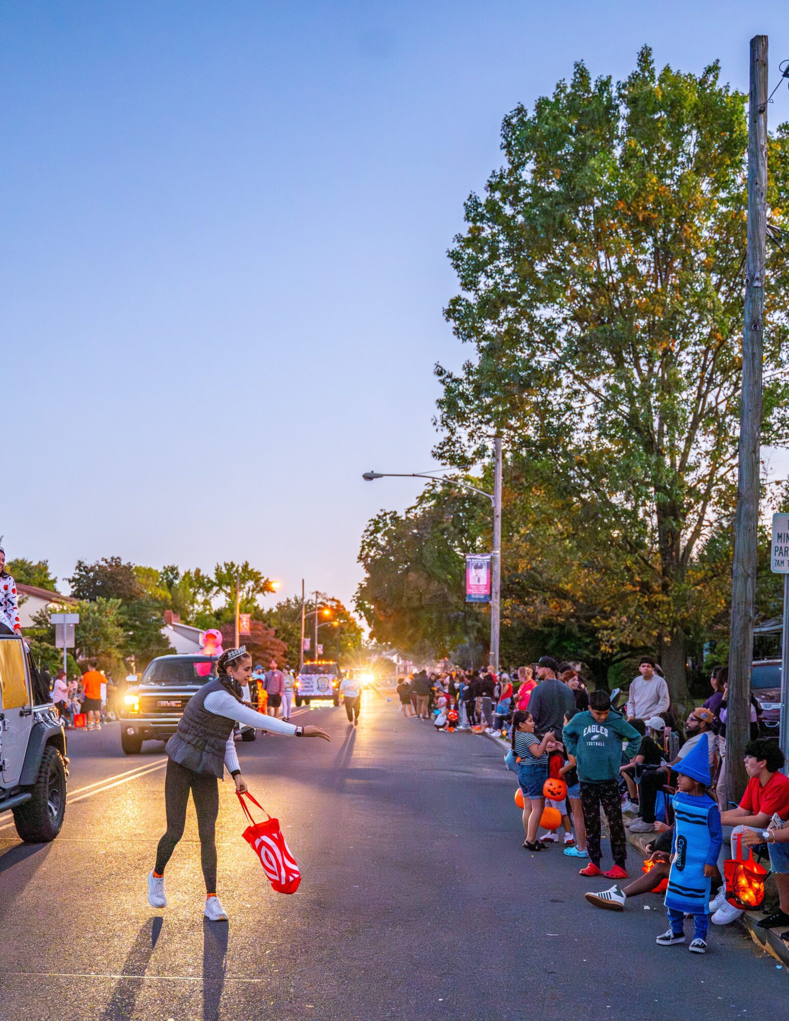 People line up along the sidewalk, children and adults, with trees and parked cars nearby, during daytime.