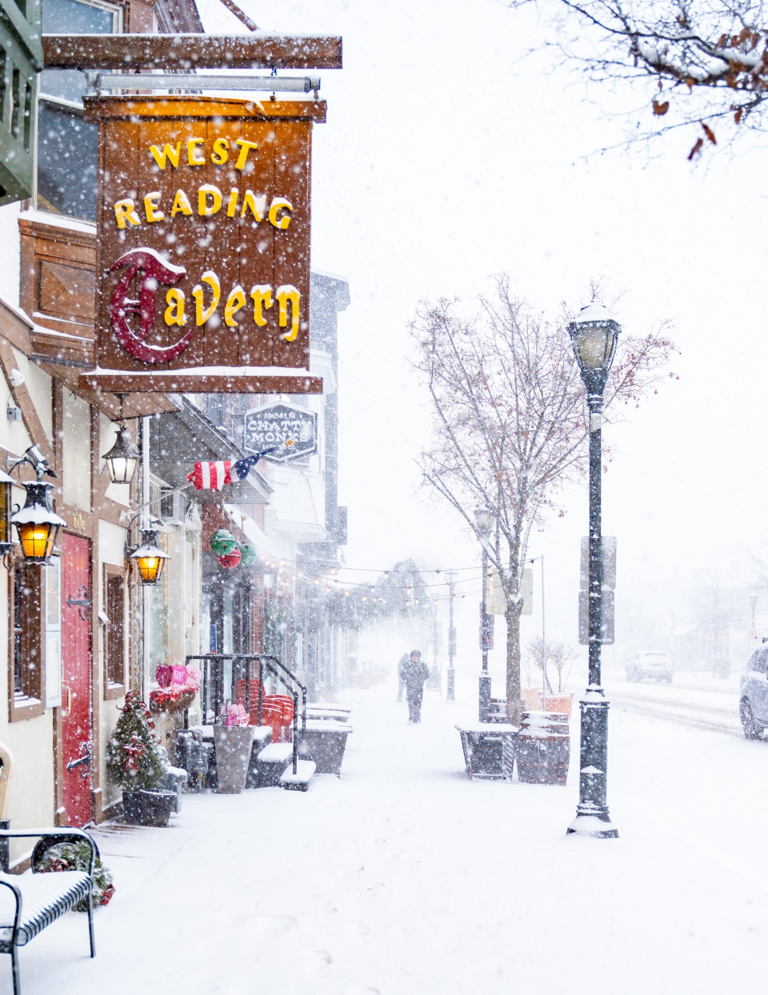 Snow-covered street with storefronts, streetlights, and a person walking in the distance during snowfall.