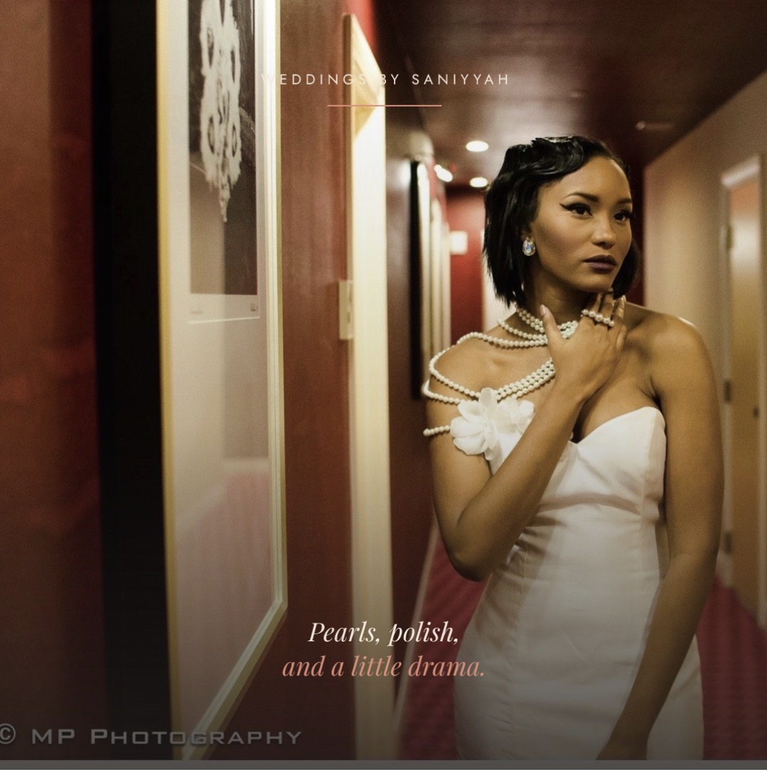 Woman in white dress with pearl necklace in hallway, holding her shoulder, looking to the side.