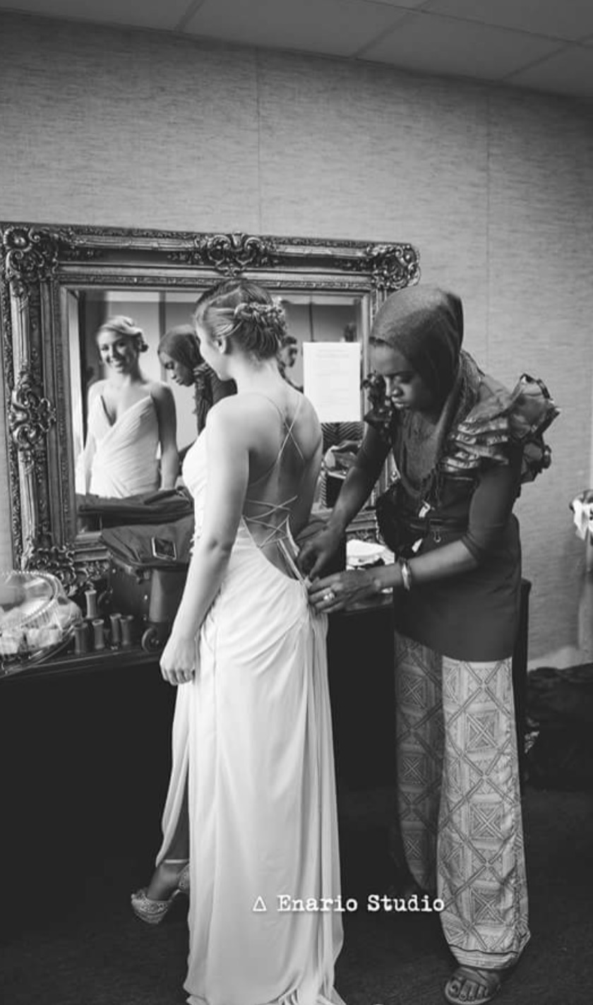 A bride in a wedding dress stands in front of a mirror, while a woman adjusts her dress. The room has a decorative mirror.