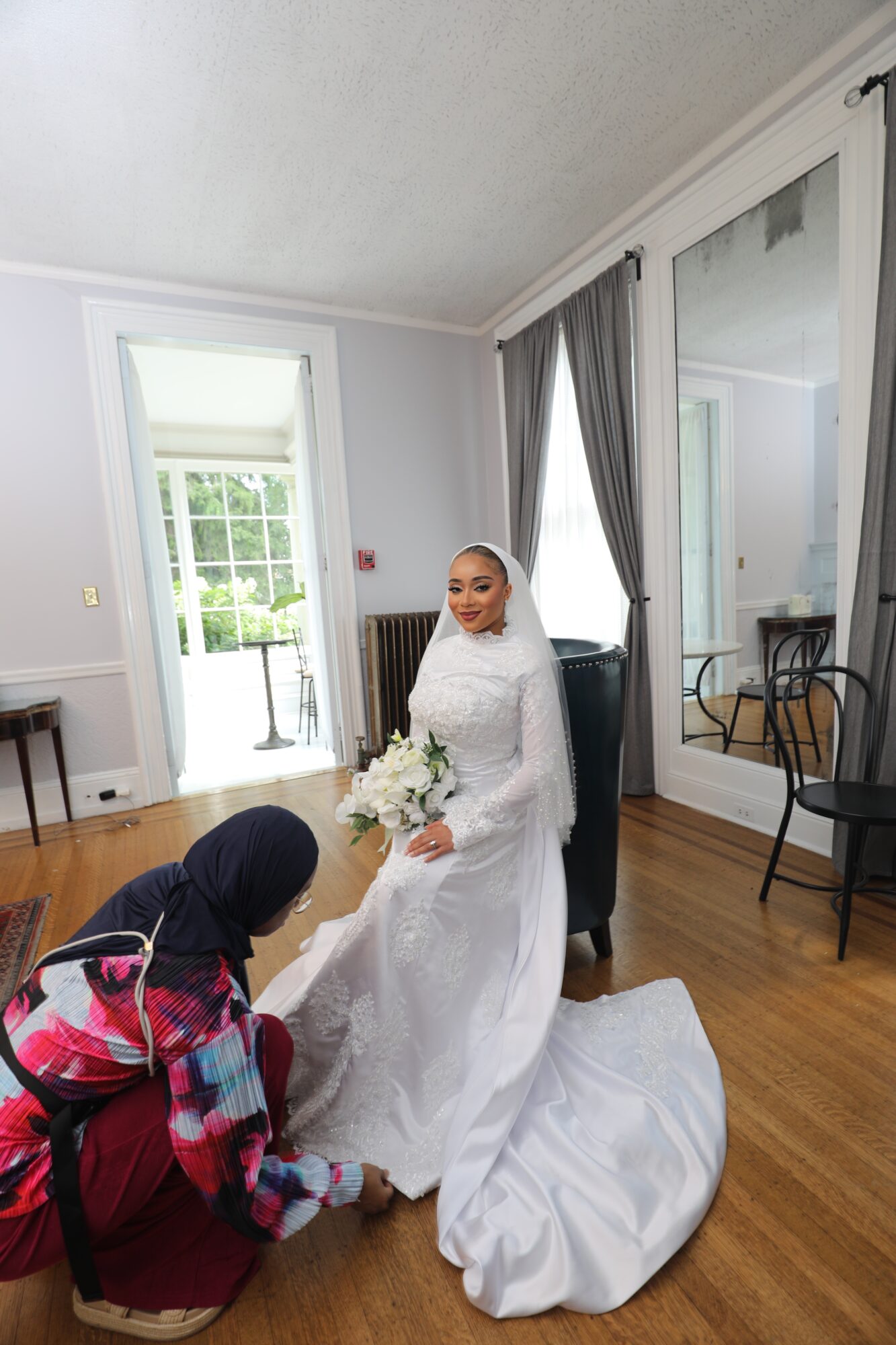 Bride in white dress holding bouquet, seated indoors with a person kneeling in front, in a room with large mirror and curtains.