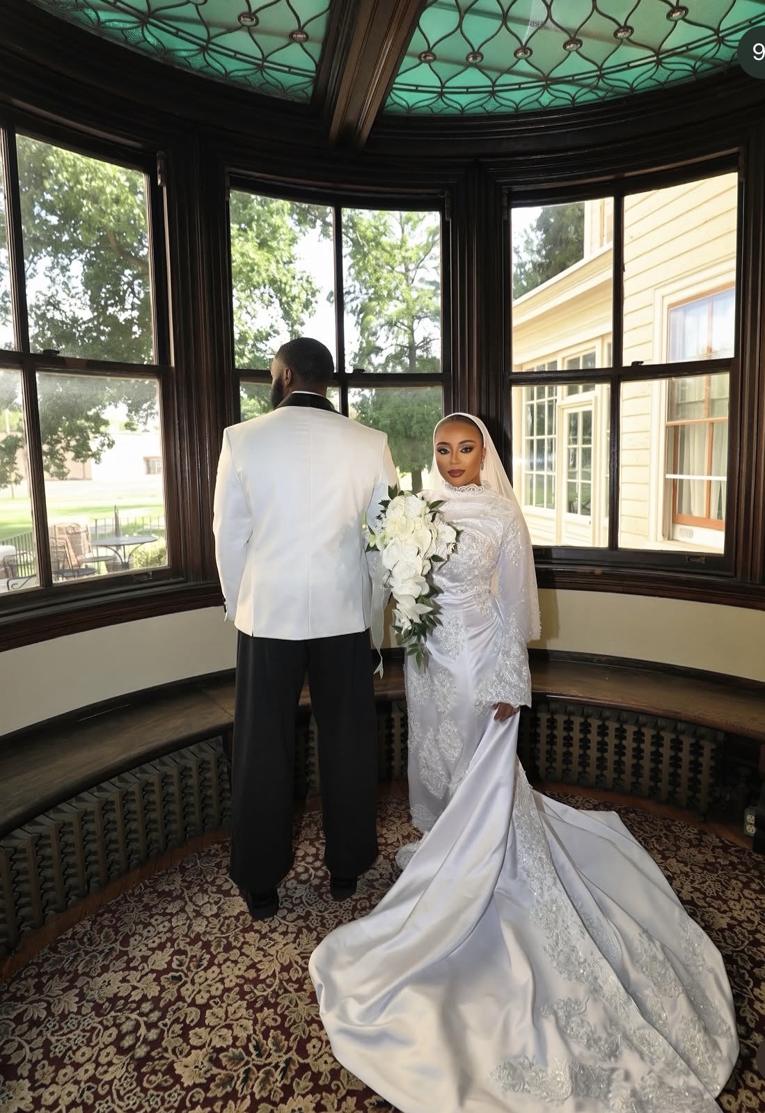Bride and groom stand inside a bay window, with trees and a house visible outside, bride holding a bouquet, bride in white gown.