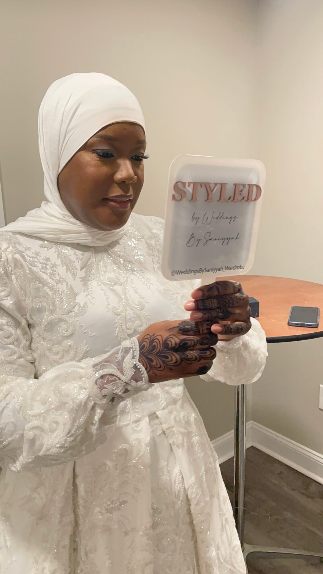 Woman in white dress and headscarf looking at a mirror, holding a small object, standing in a room with a table.