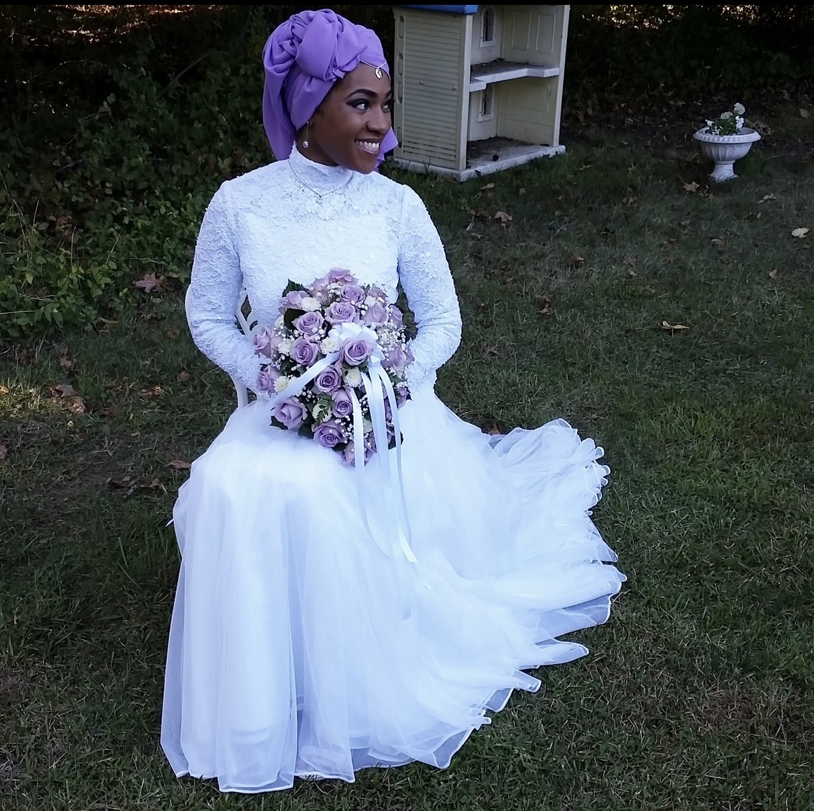 Woman in white dress holding a bouquet of purple and white flowers, sitting outdoors on grass, smiling, wearing a purple headwrap.
