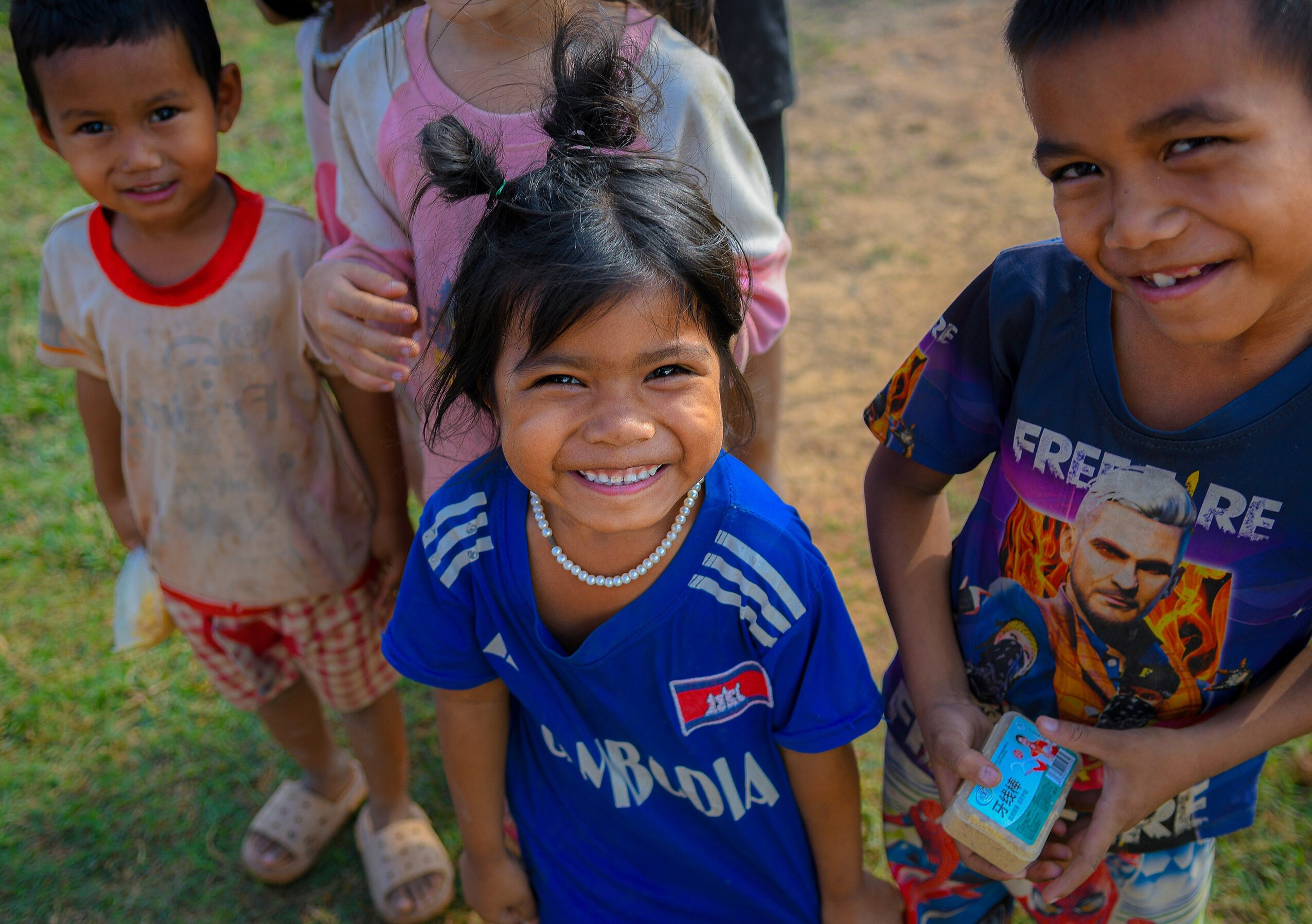 Group of children smiling outdoors, girl in blue shirt with Uganda flag, others around her, sunny day.
