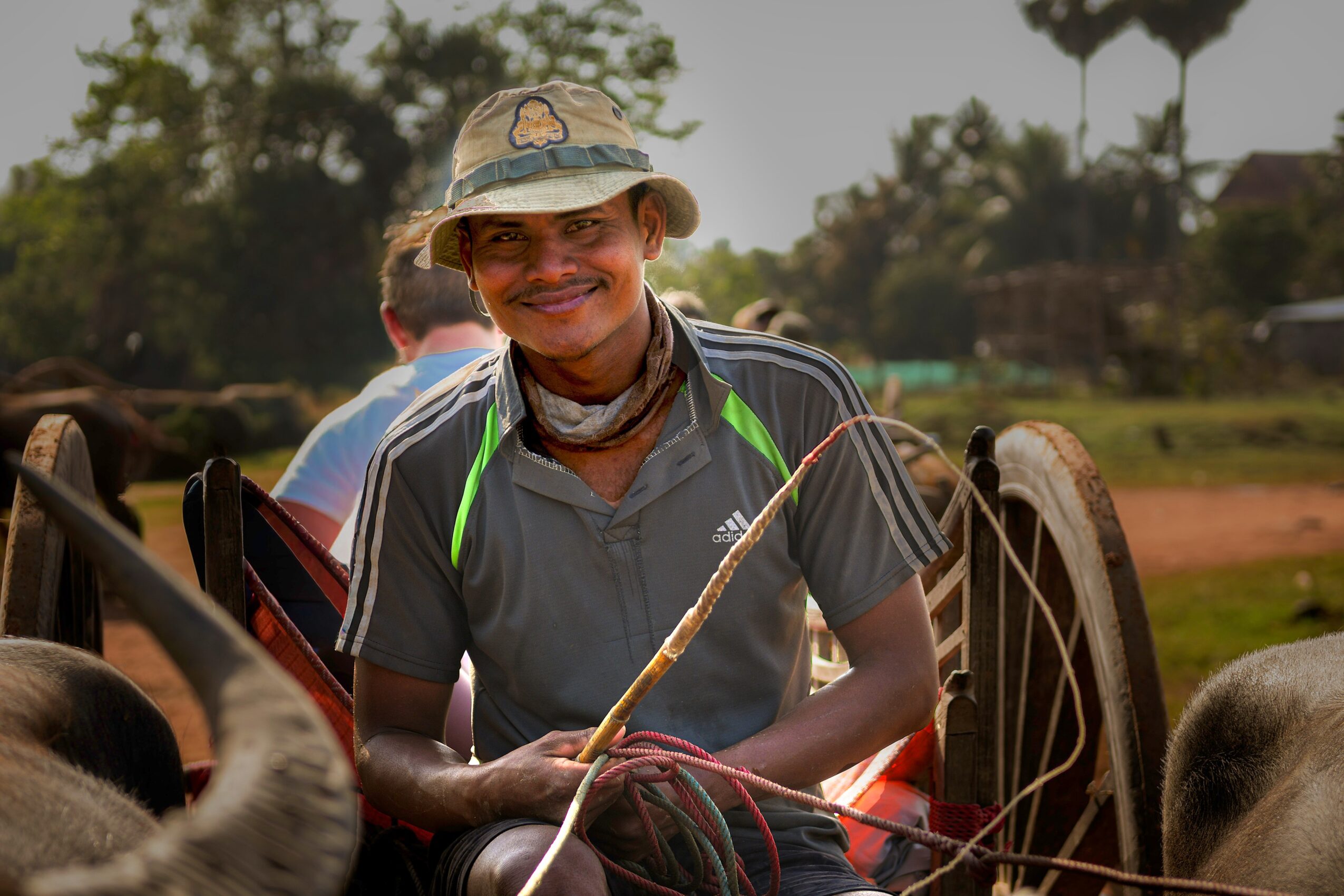 Smiling person sitting on a cart outdoors, holding a stick, wearing a hat and casual clothing.