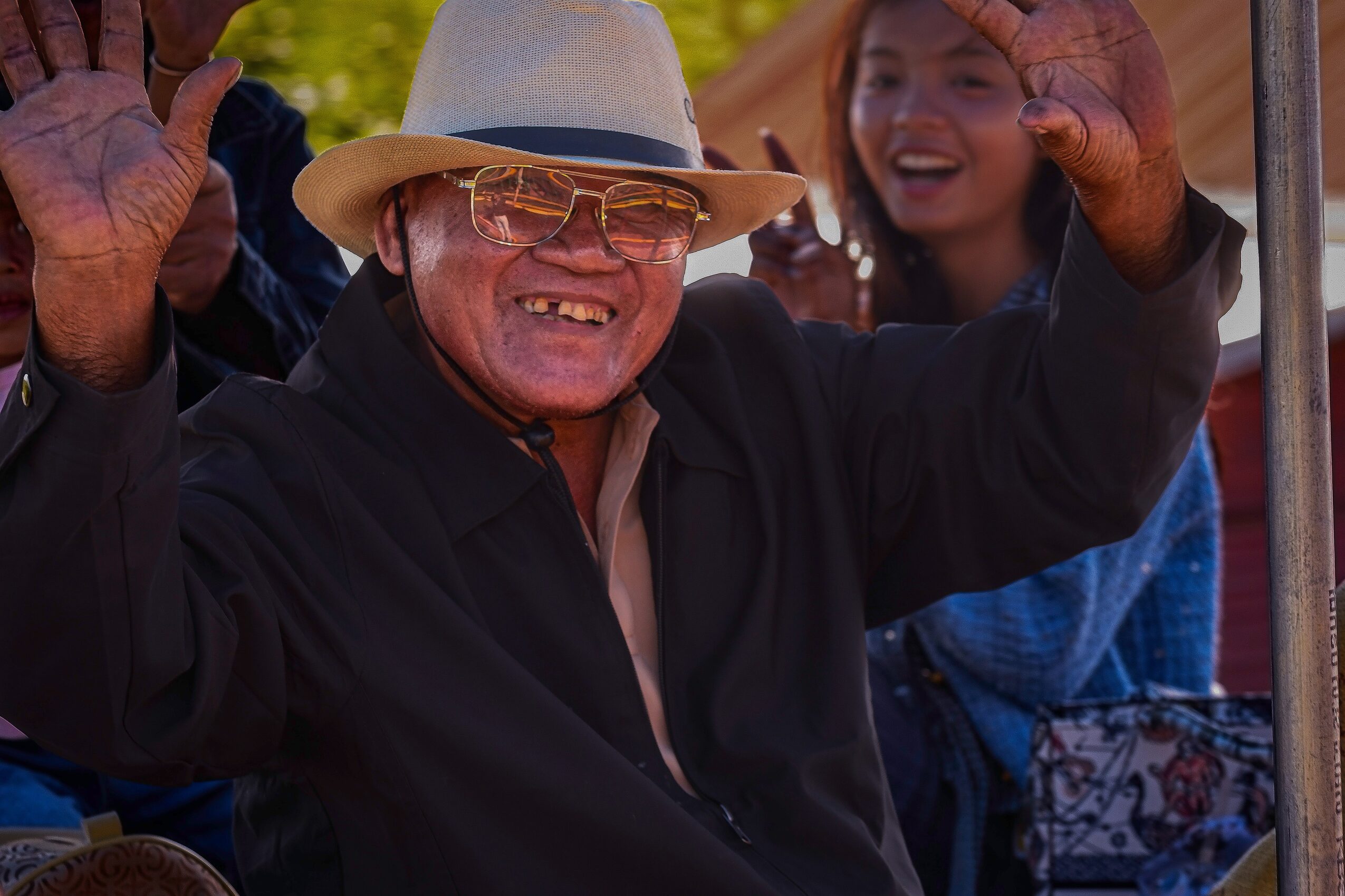 Older man wearing glasses, a hat, and a dark jacket, smiling and waving with a young woman in the background.
