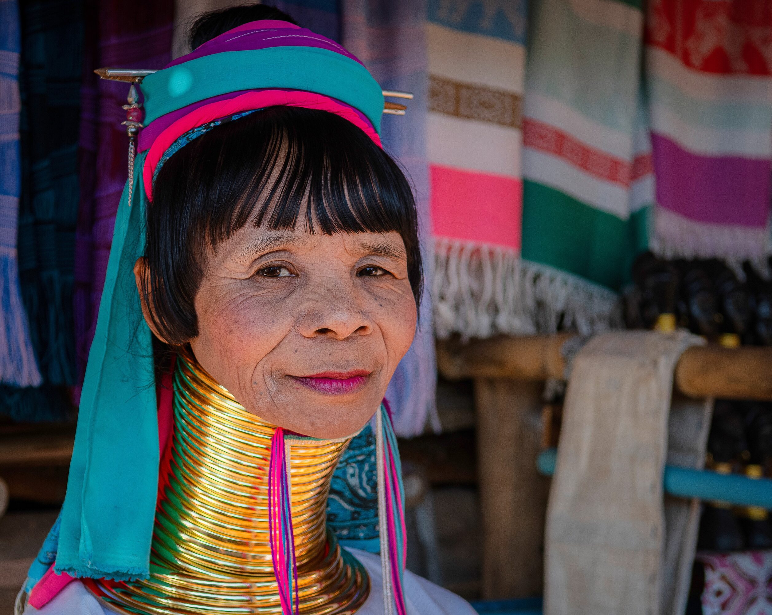 Woman wearing traditional jewelry and colorful clothing, with textiles hanging in the background.