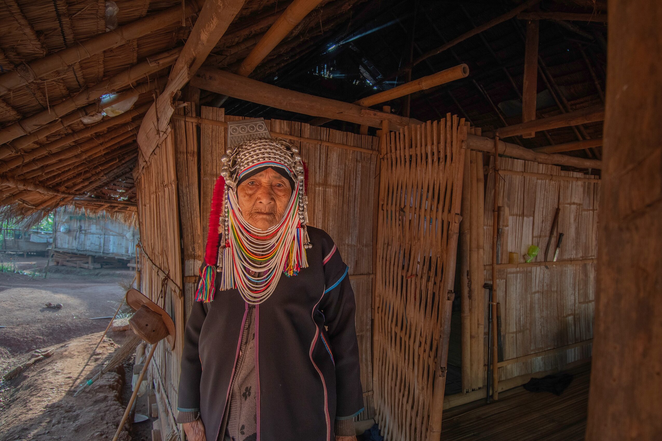Woman wearing traditional jewelry and headscarf standing in front of a bamboo fence, sunlight casting shadows.
