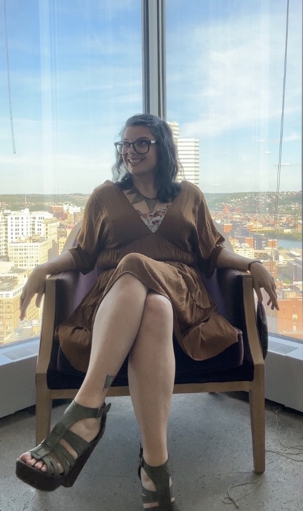 Woman with glasses sitting on a chair in front of a large window with city view, smiling, wearing a brown dress and sandals.