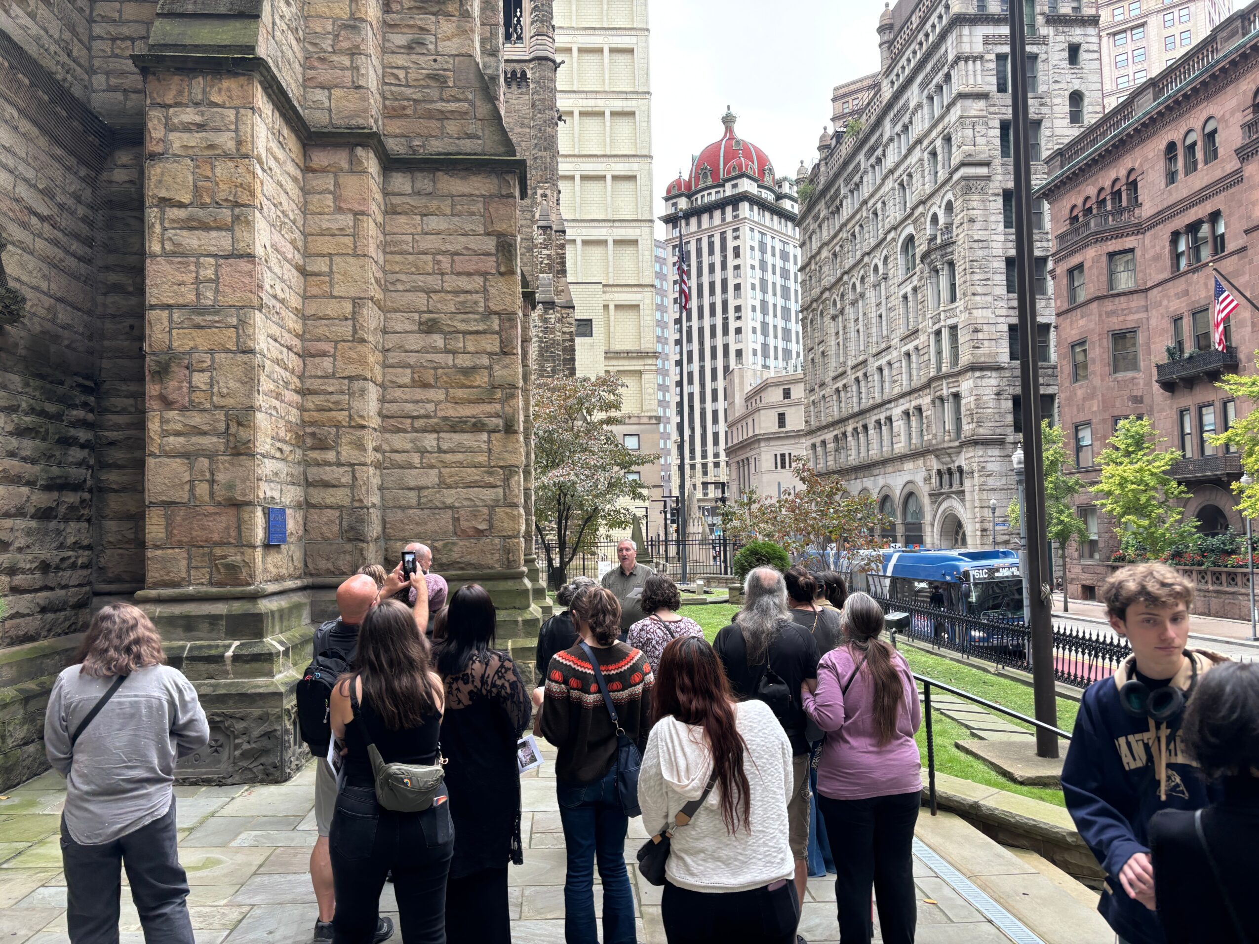 Group of people standing on a city sidewalk near historic buildings, with tall skyscrapers in the background.