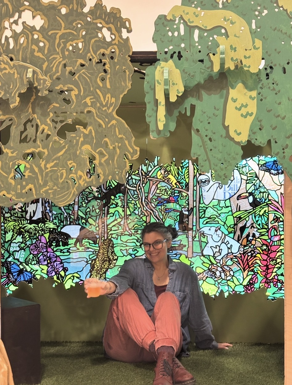 Person sitting on the floor in front of a colorful, illustrated forest background, smiling, wearing glasses, a denim jacket, and pink pants.