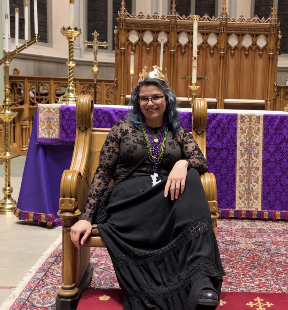 Woman sitting on a wooden throne-like chair in a church, wearing a crown and black dress, smiling, with purple and gold decorations behind her.