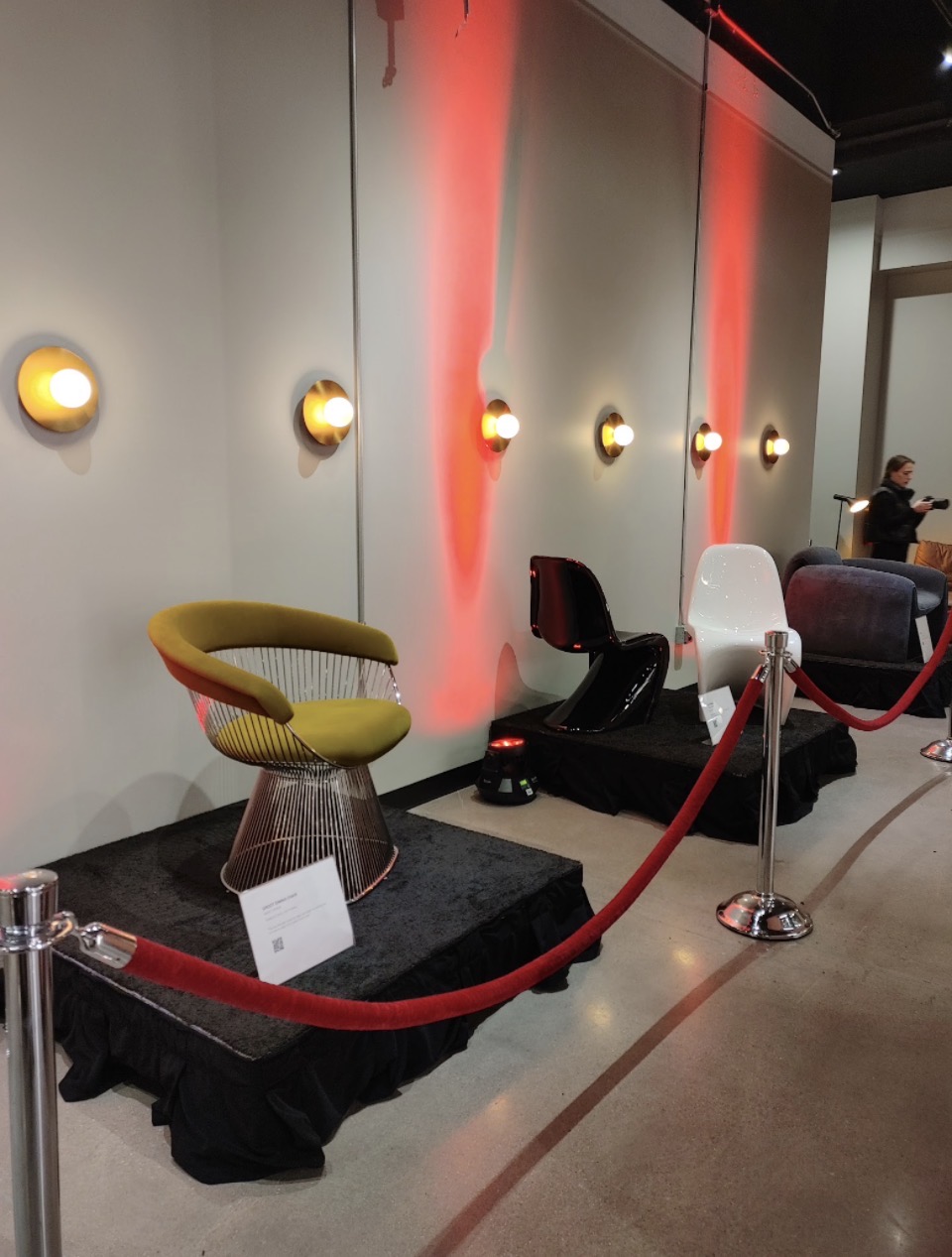 Three chairs displayed on black platforms with red velvet ropes, wall with lights and red lighting effects in background.
