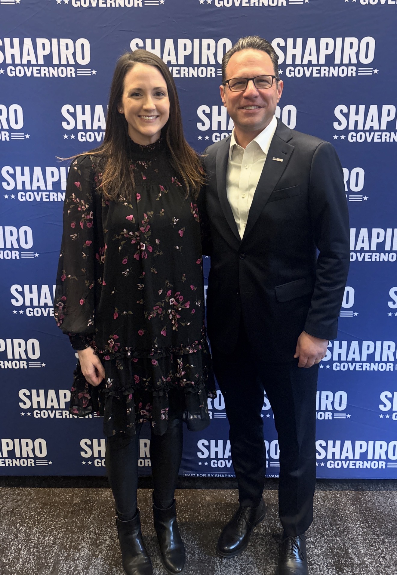 Two people standing in front of a blue backdrop with white text, smiling, one woman and one man, both dressed professionally.