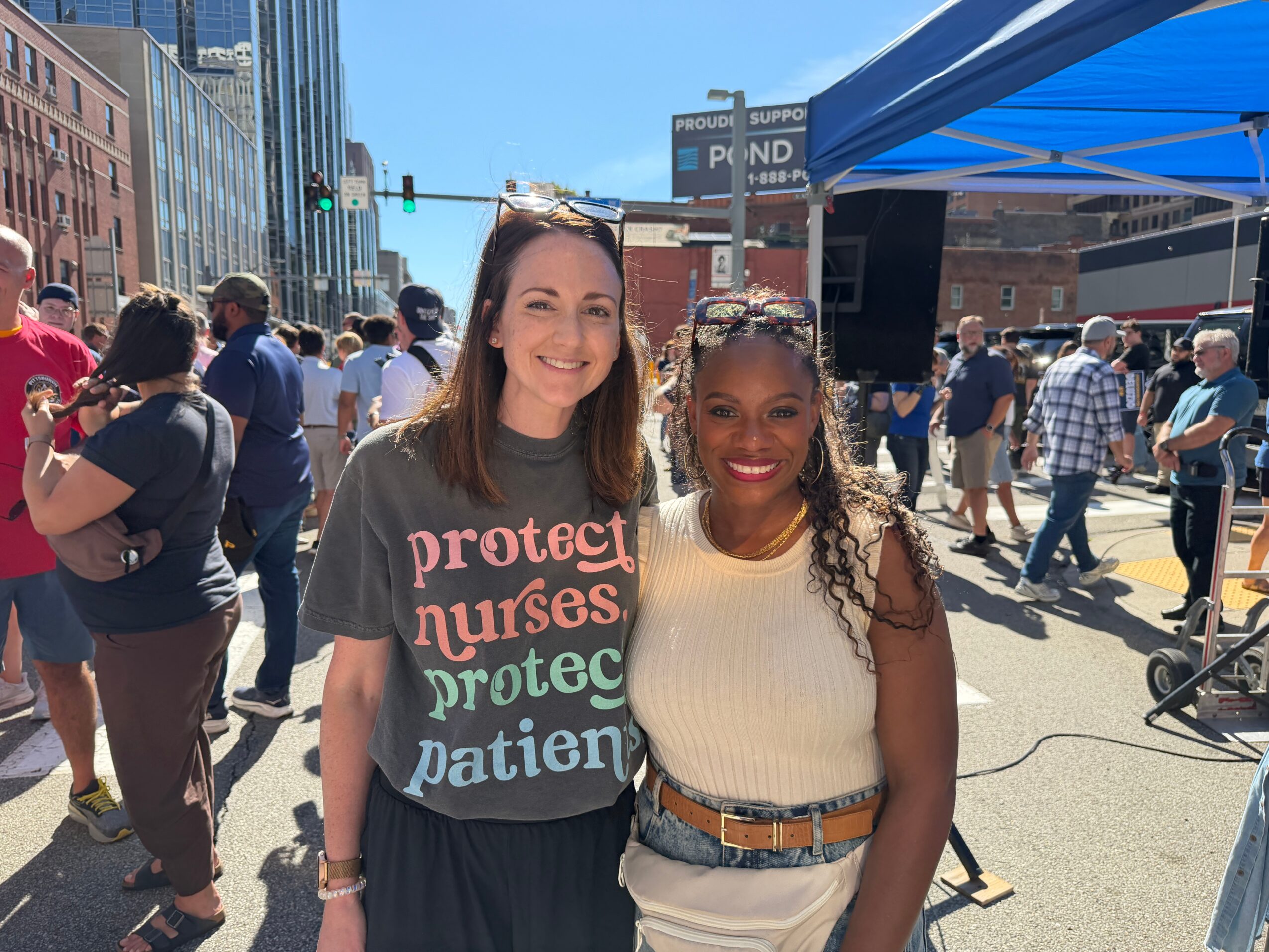 Two women smiling at an outdoor event with people and buildings in the background.