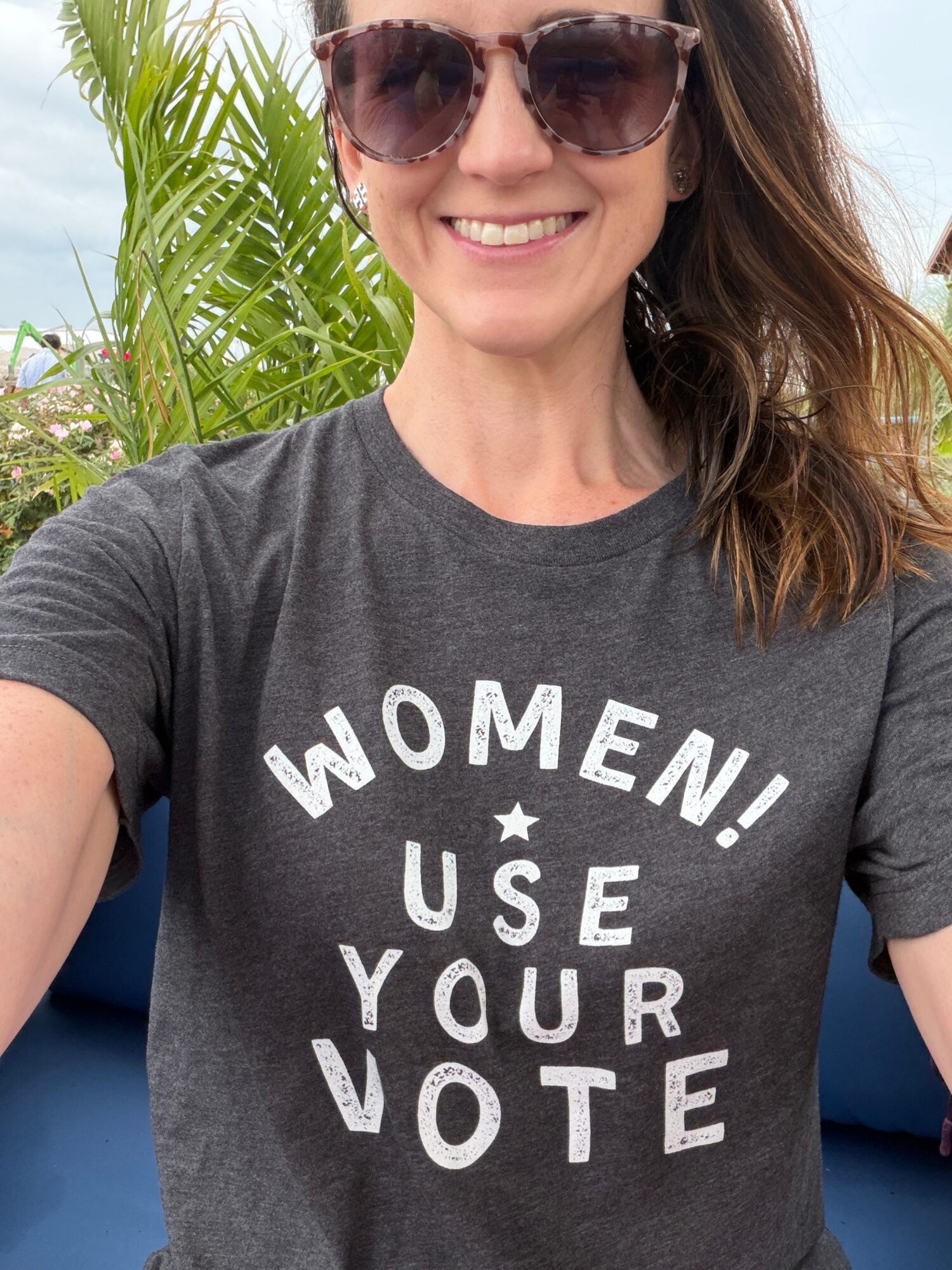Woman wearing sunglasses and a gray T-shirt with text, smiling outdoors with green plants in background.