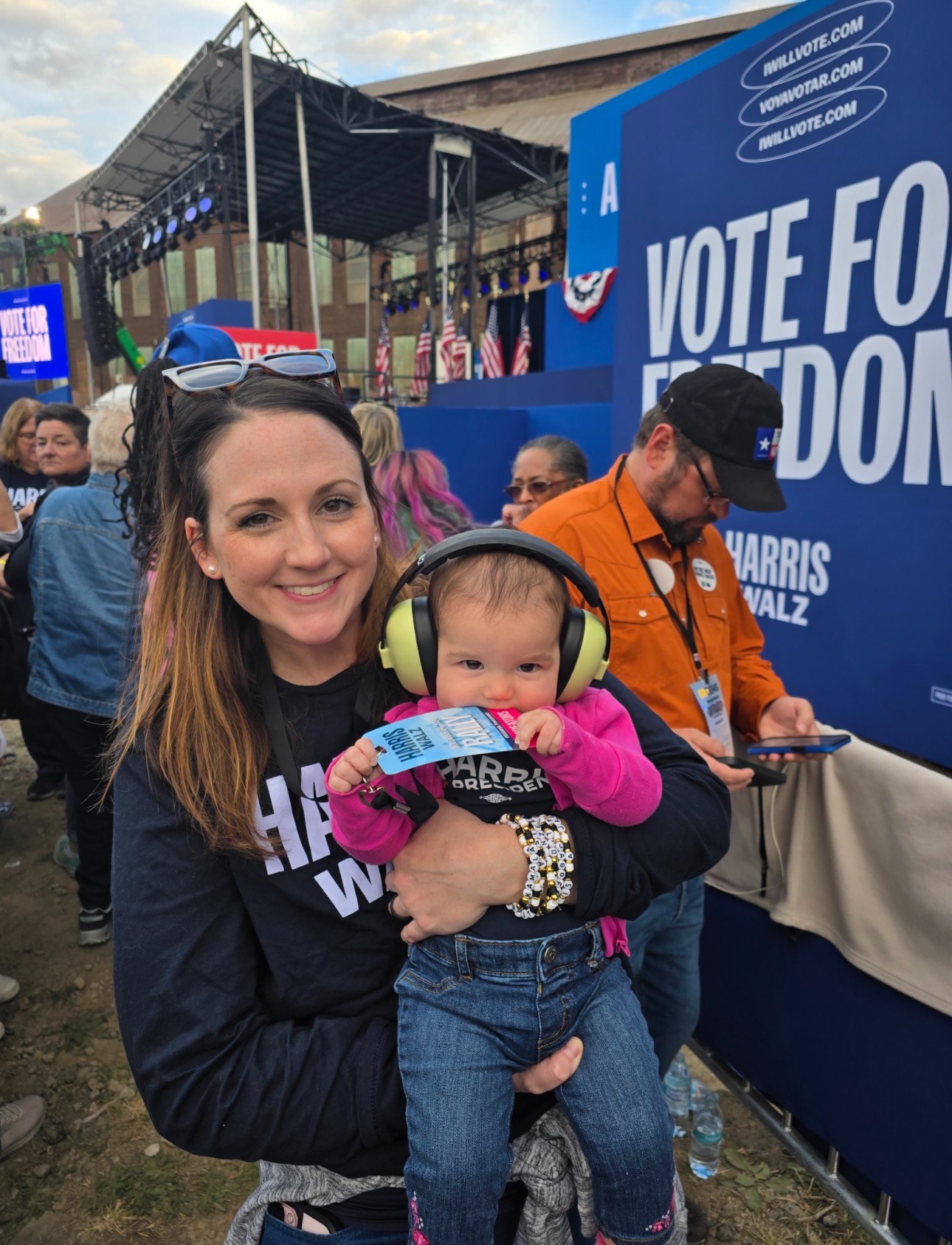 Woman holding a baby at an outdoor event with a voting sign and stage in background.