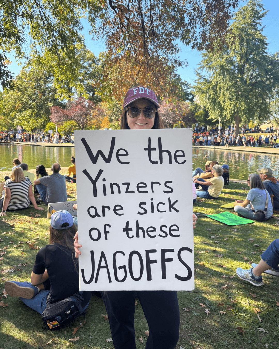 Person holding a sign protesting Yinzers at a park with trees and people sitting on grass.