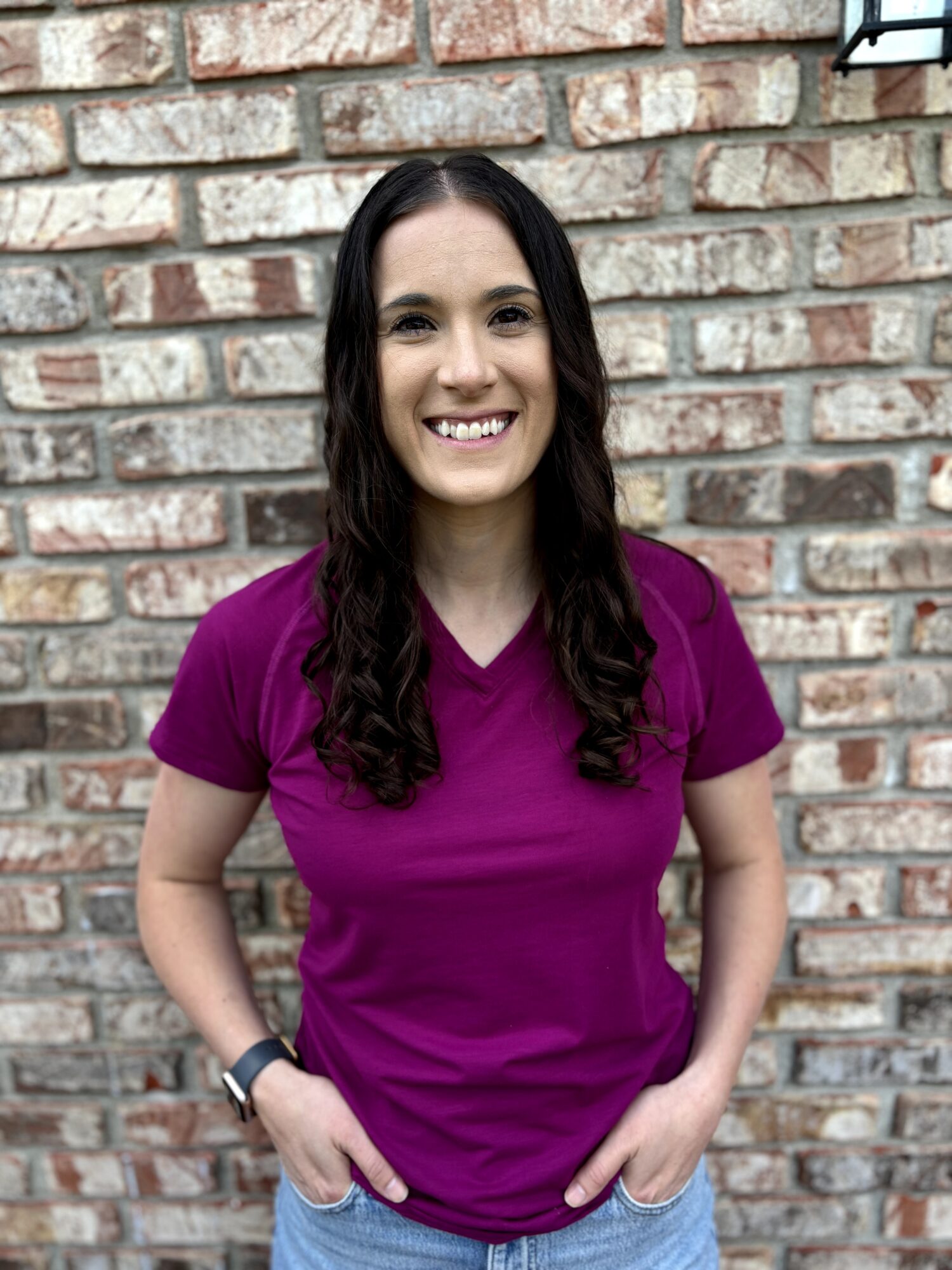 Young woman with long dark hair, smiling, wearing a purple shirt, standing in front of a brick wall.