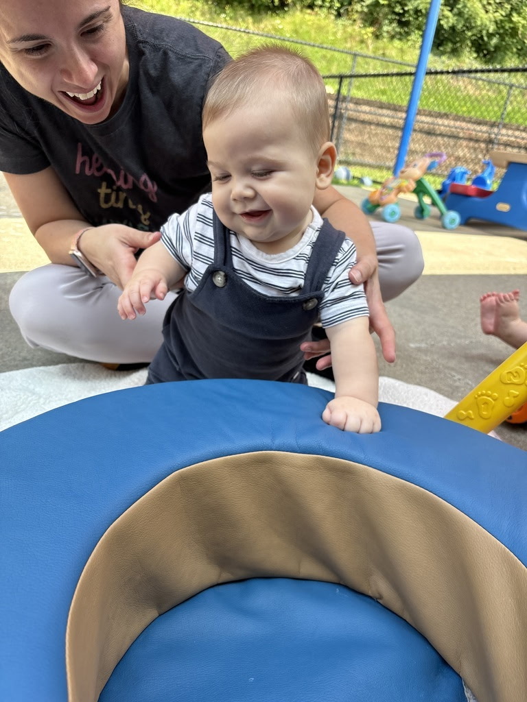 A smiling woman and a young child playing outdoors near a blue and yellow play structure, with toys in the background.
