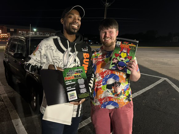 Two men standing outdoors at night, smiling, holding comic books, with a car and parking lot in background.