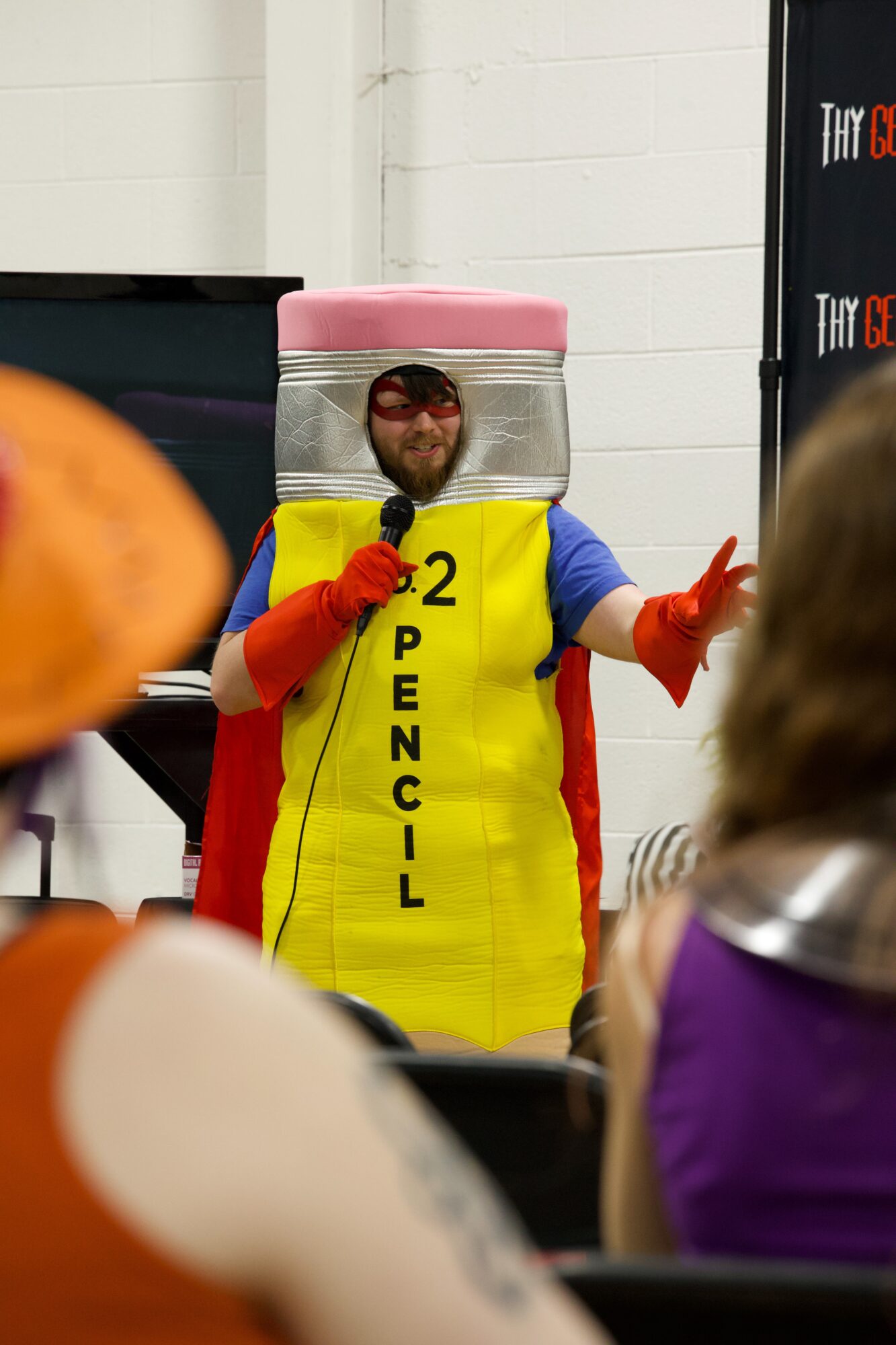 Person dressed as a pencil with a pink eraser head, yellow body, blue arms, red cape, and sunglasses, speaking to an audience.