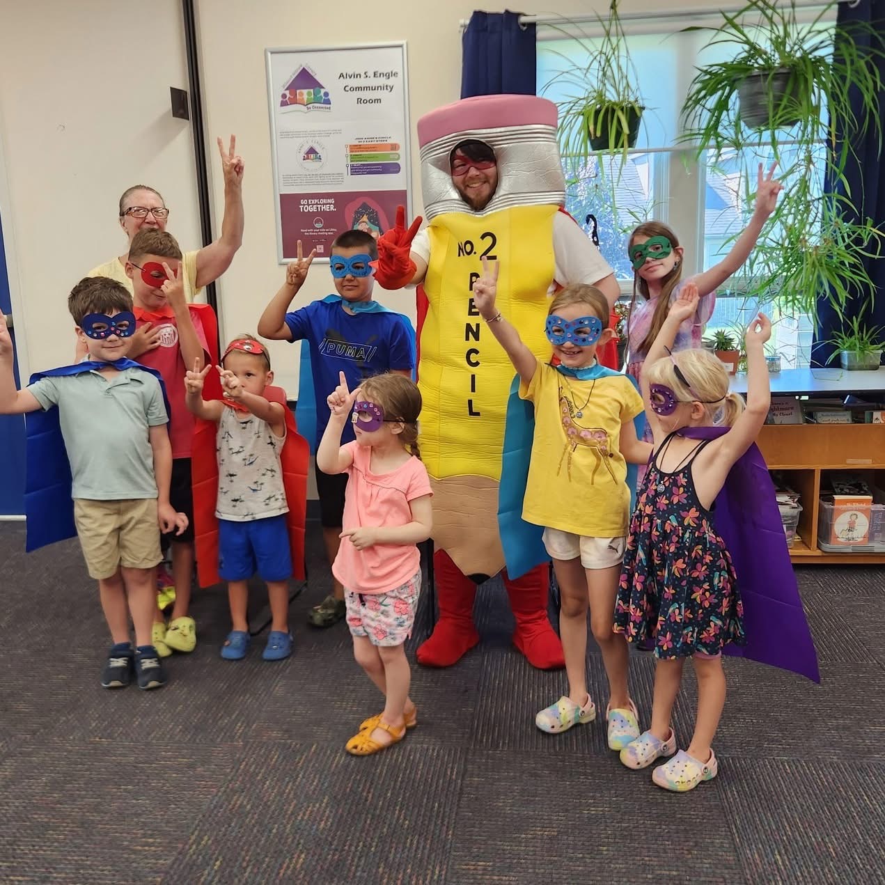 Group of children and adults with a person in a large pencil costume, all wearing masks, in a classroom setting.