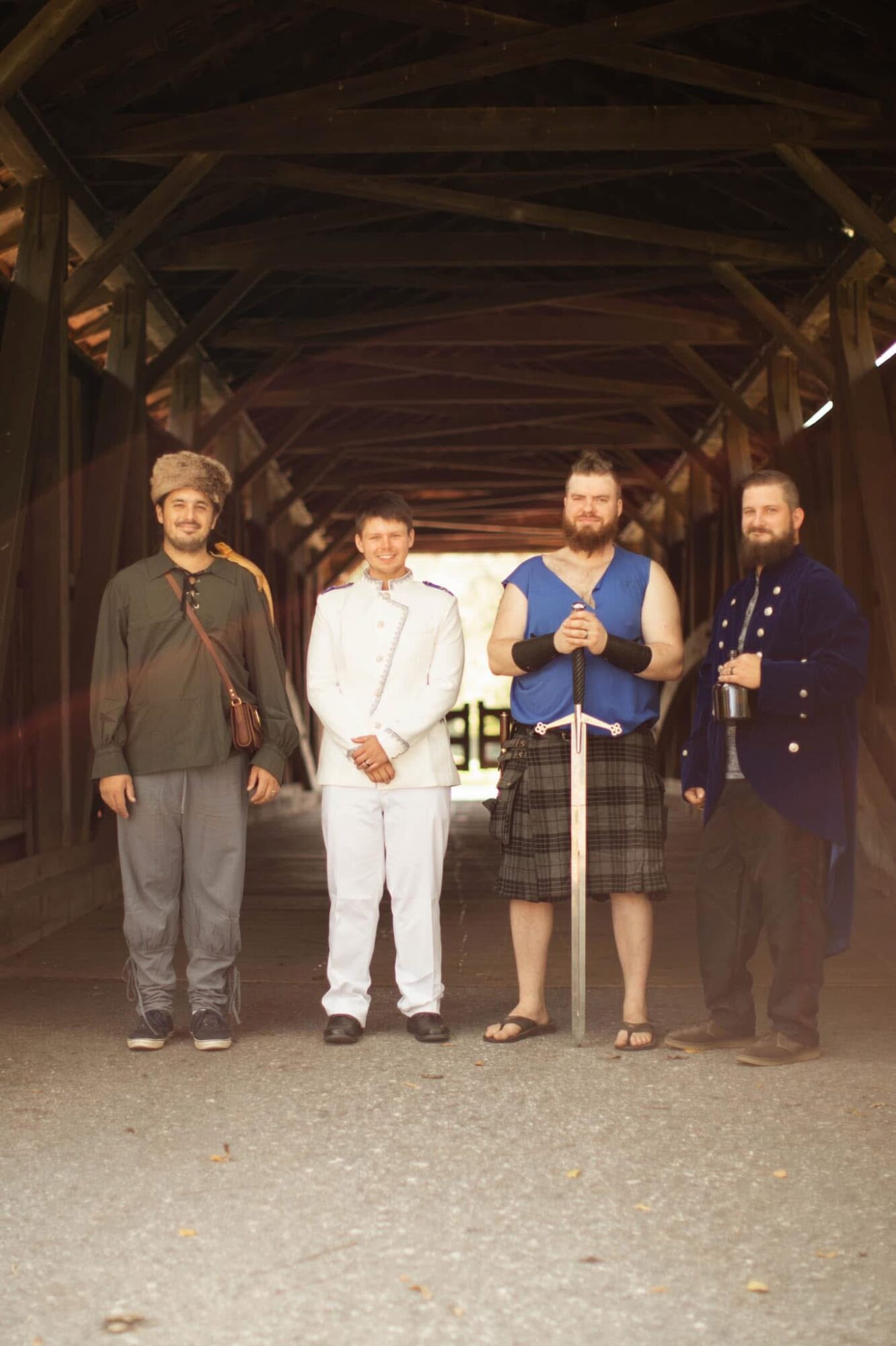 Four men stand under a wooden bridge, dressed in casual and formal clothing, with a bright background behind them.