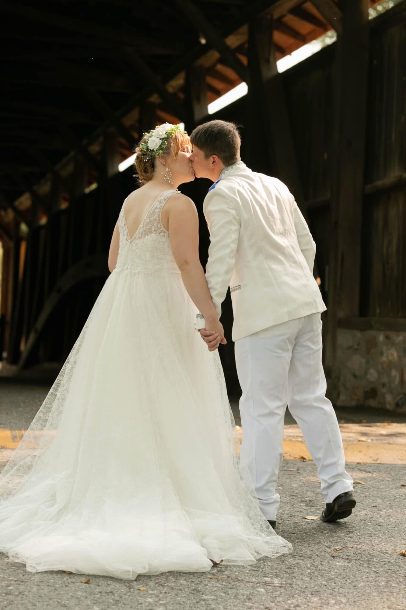 A bride and groom hold hands and kiss outdoors near a wooden structure, with the bride in a white gown and floral headpiece.