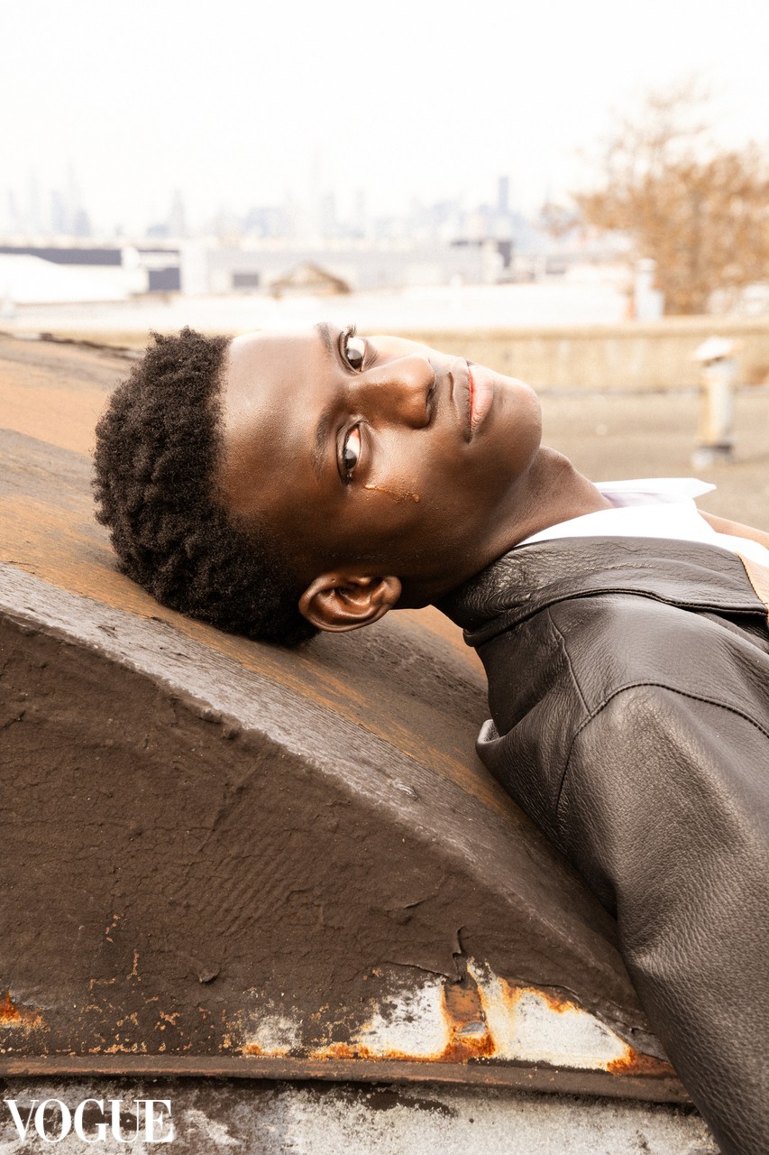 Young person with short curly hair lying on a sloped surface outdoors, wearing a black leather jacket, looking at the camera.