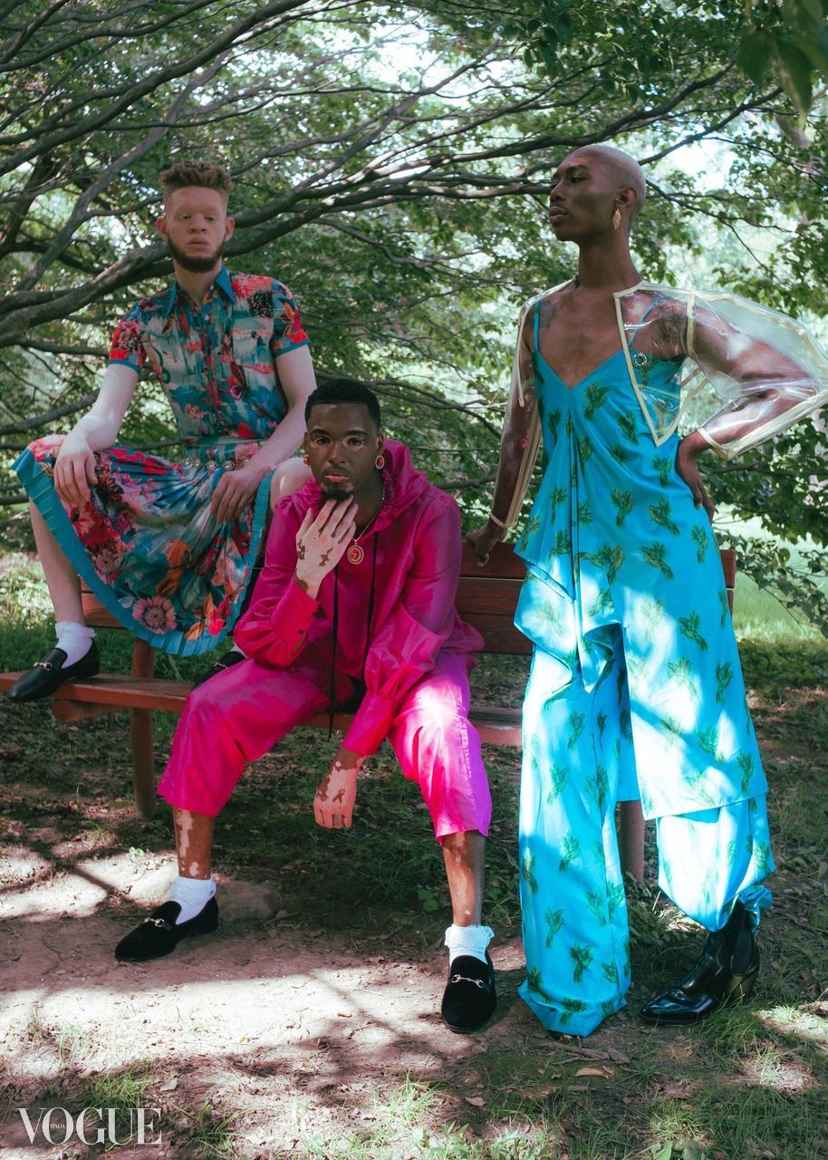 Three people outdoors under trees, one sitting, one standing, one kneeling, wearing colorful clothing, with sunlight filtering through leaves.
