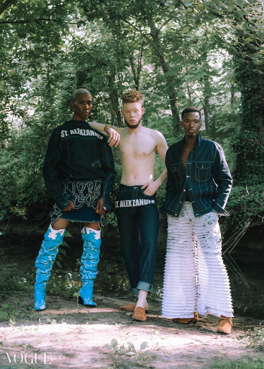 Three people standing outdoors in a wooded area, two wearing patterned pants and one shirtless, with trees in the background.