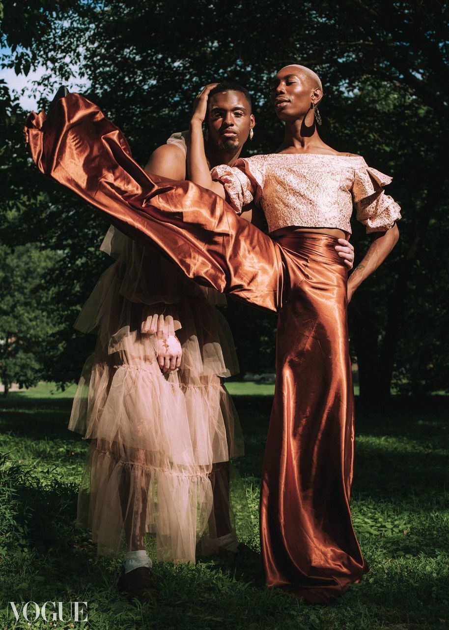 Two women in fashionable outfits standing outdoors with trees in the background, one with a flowing skirt and the other with high-waisted pants.