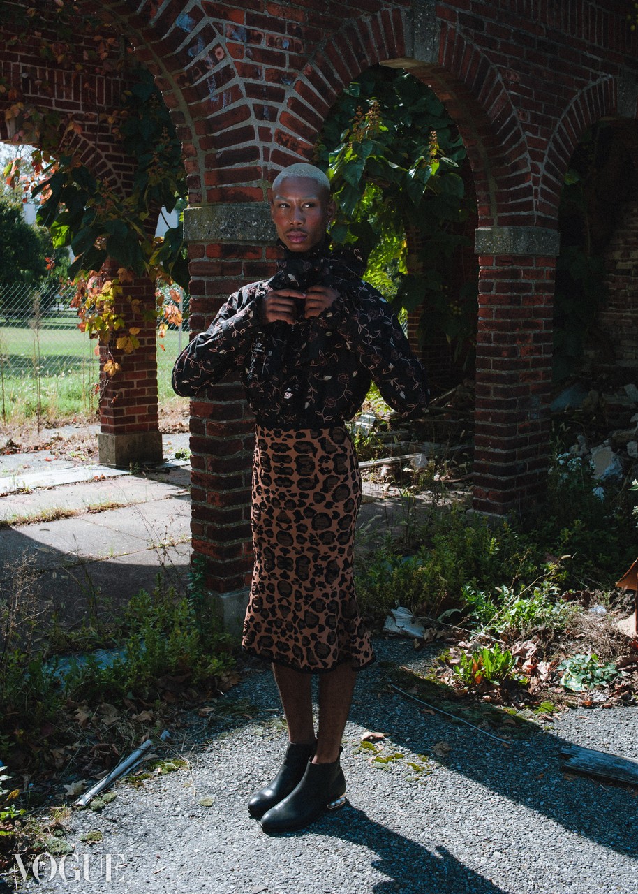 Person with short hair taking a mirror selfie outdoors, wearing a leopard print skirt and black top, standing under brick arches.