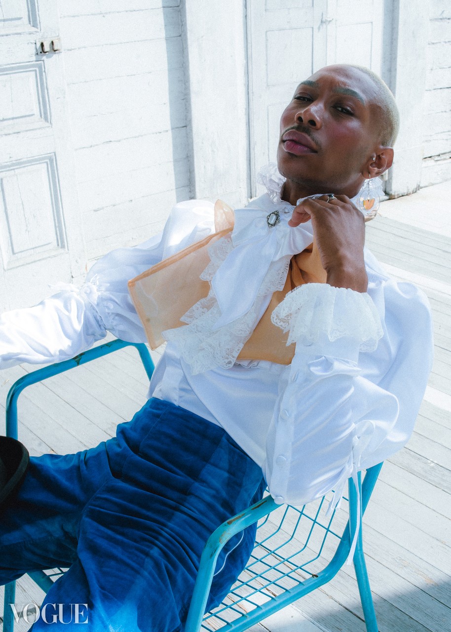 Person with short hair and earrings, wearing a white ruffled blouse and blue pants, sitting on a blue chair outdoors.