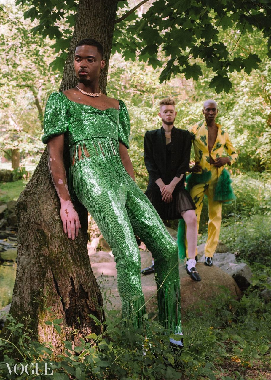 Three people outdoors among trees, one in a green dress leaning against a tree, two others standing behind, one in black, one in yellow.