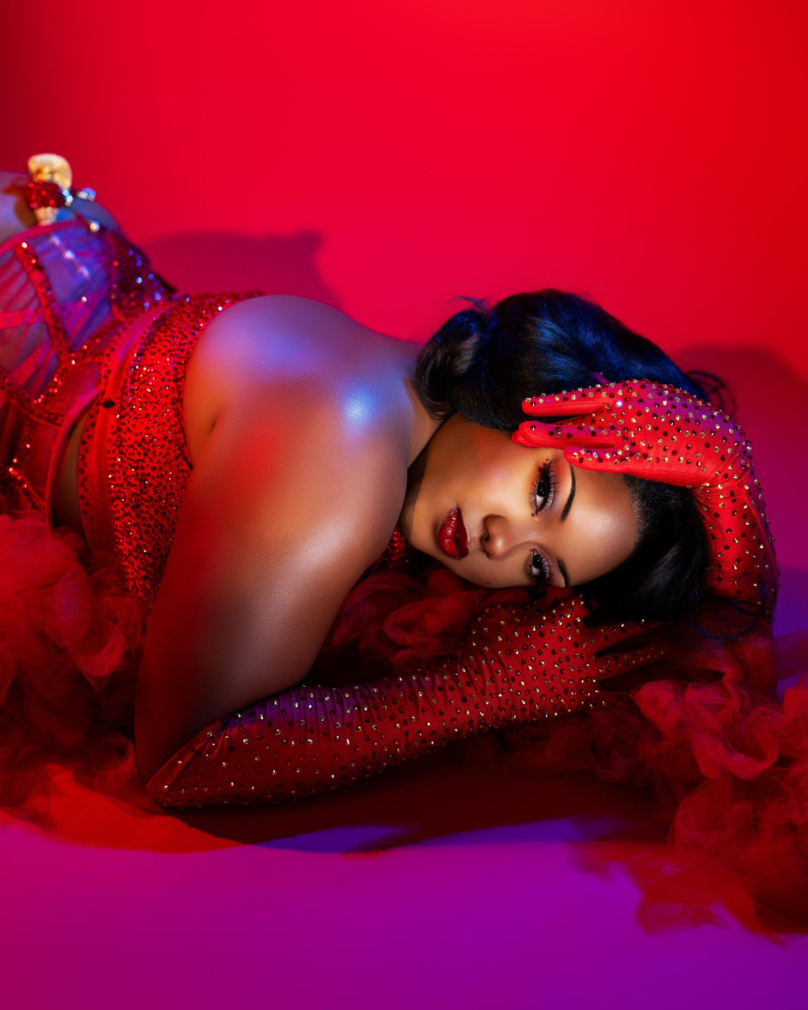 Woman lying on red fabric, wearing red gloves and dress, with dark hair and makeup, against a red background.