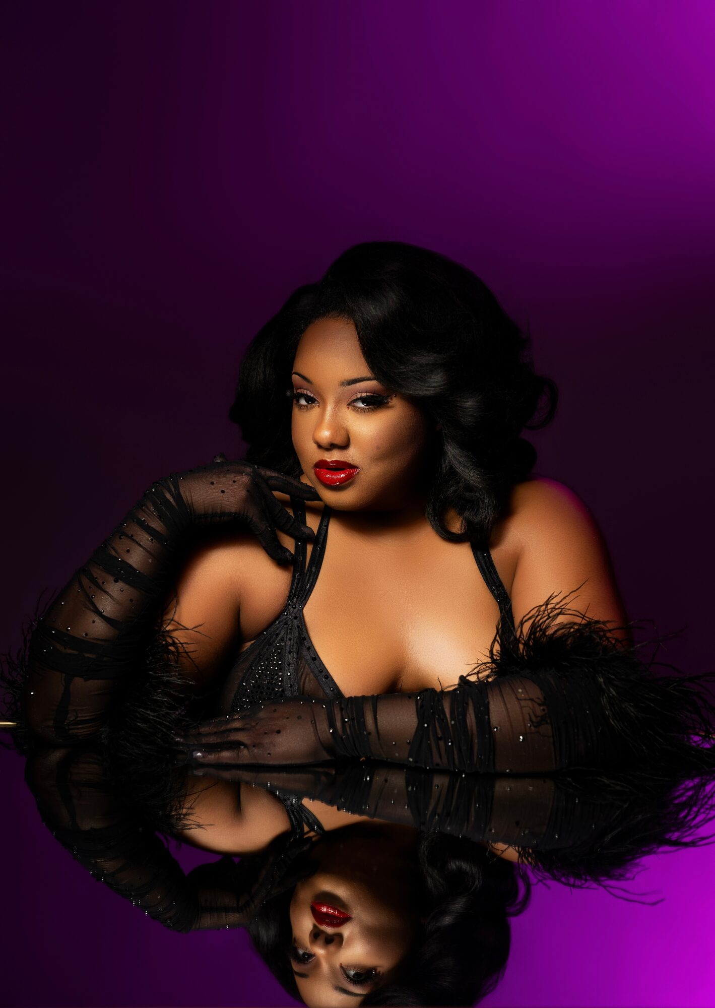 Woman with dark hair and makeup, wearing black lace gloves and a revealing top, posed against a purple background.
