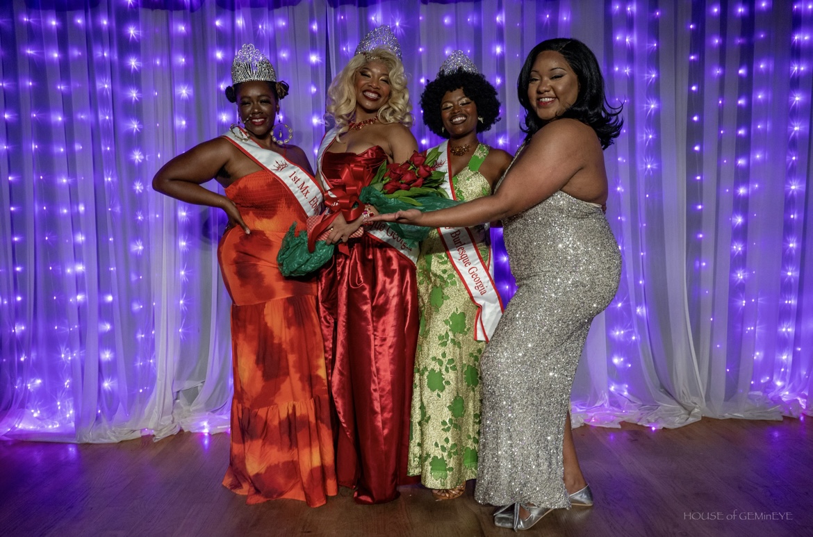 Four women in elegant dresses stand together in front of purple string lights, smiling and holding a bouquet.