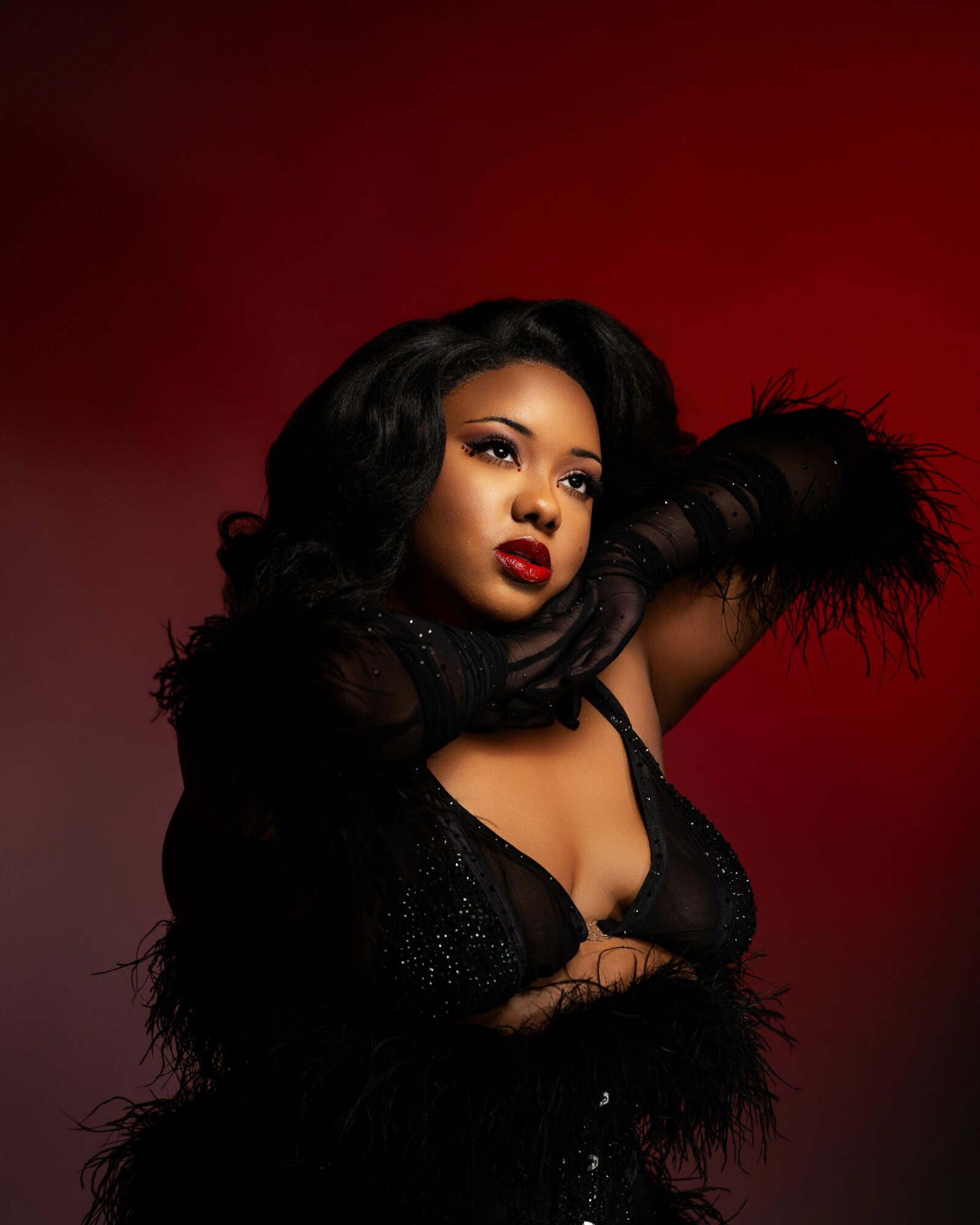 Woman with long black hair, red lipstick, and black outfit with feather details, posing with arms raised against a red background.