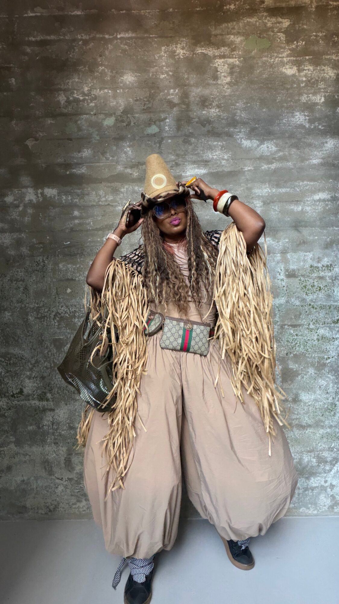 Person wearing a straw hat, sunglasses, and a fringed outfit, standing against a textured wall.