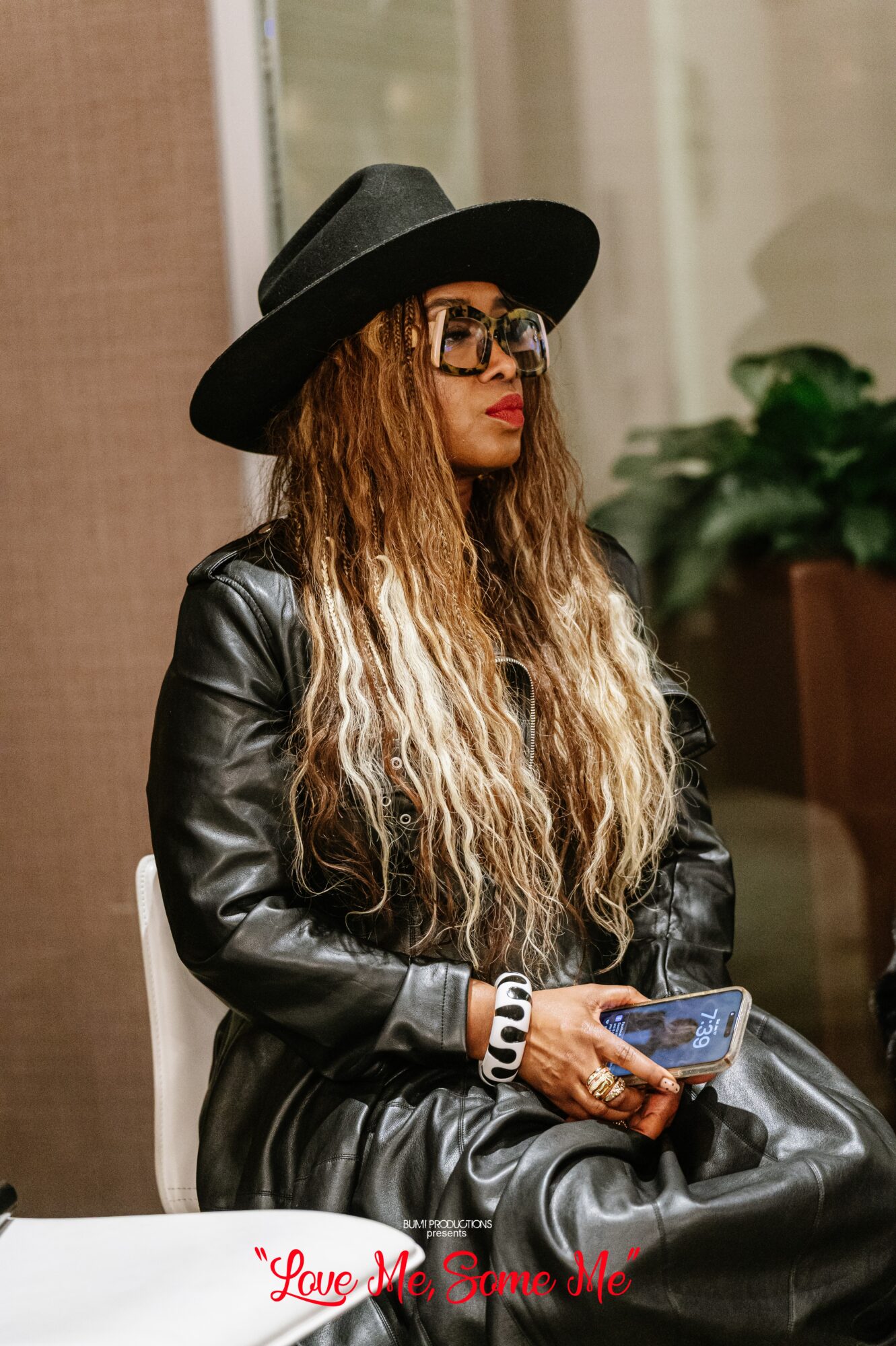 Woman with long wavy hair, wearing a black hat, sunglasses, leather jacket, holding a phone, sitting indoors.