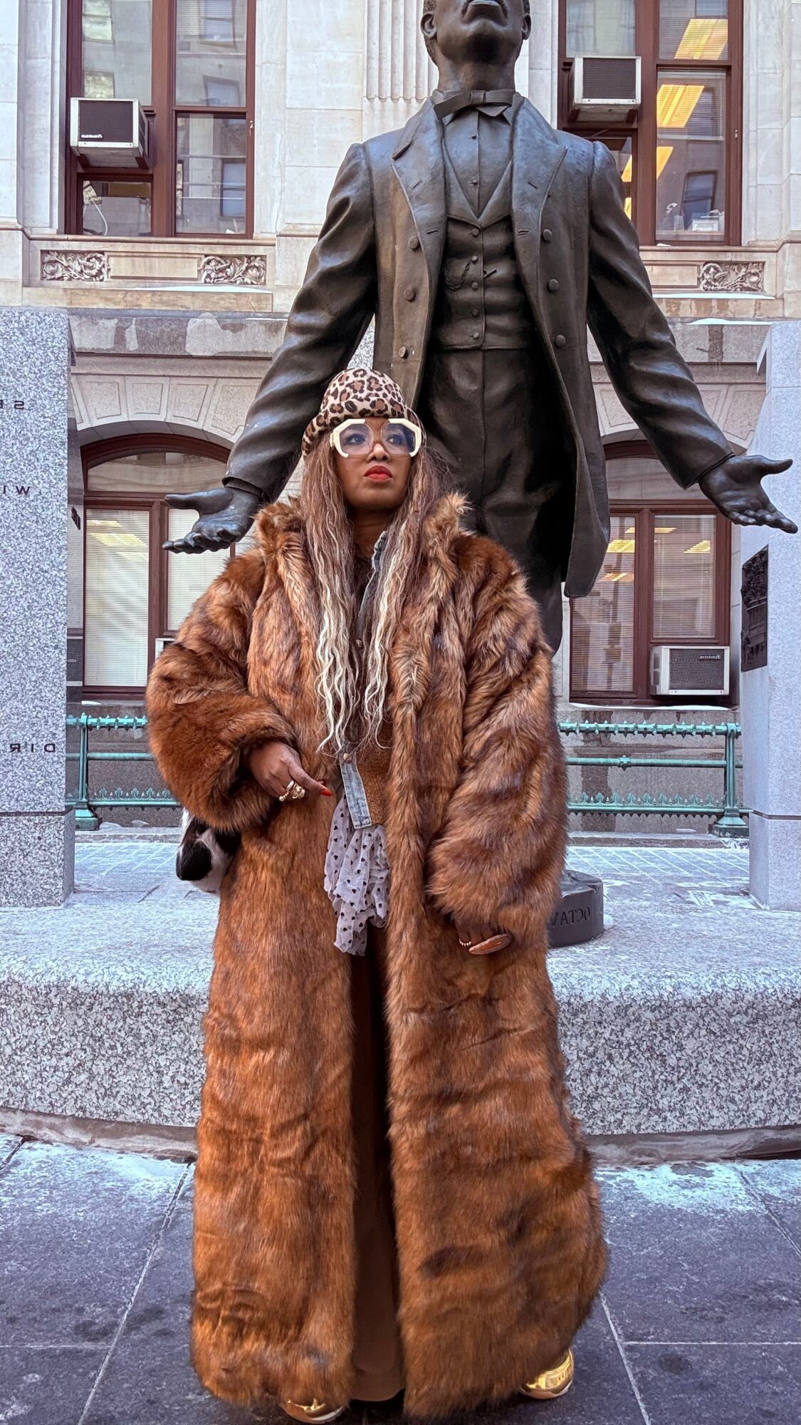 Woman in a long fur coat and sunglasses standing in front of a large statue of a man in a suit, city background.