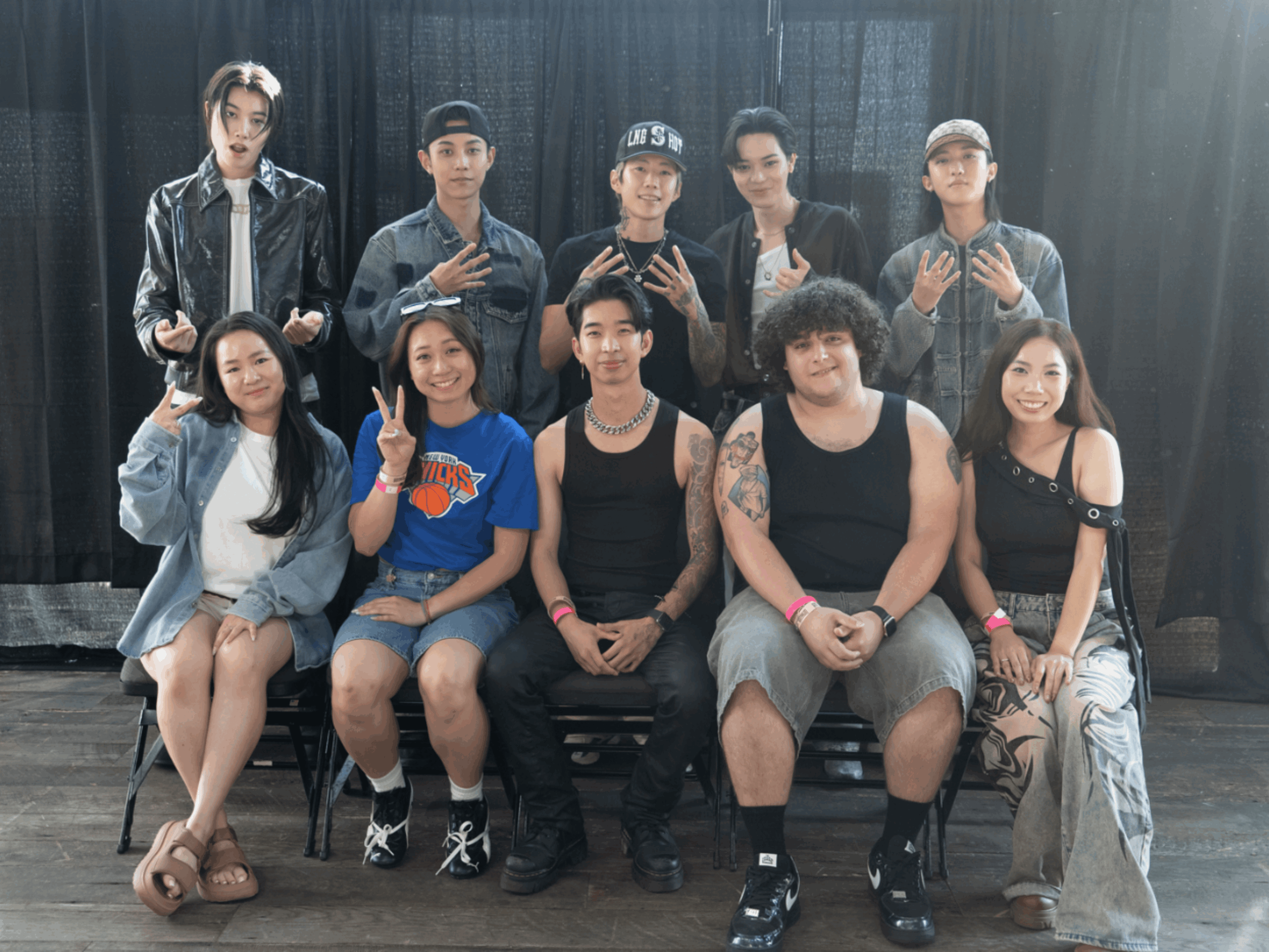 Group of ten young people posing on stage with black curtains, some making peace signs, smiling, and dressed casually.