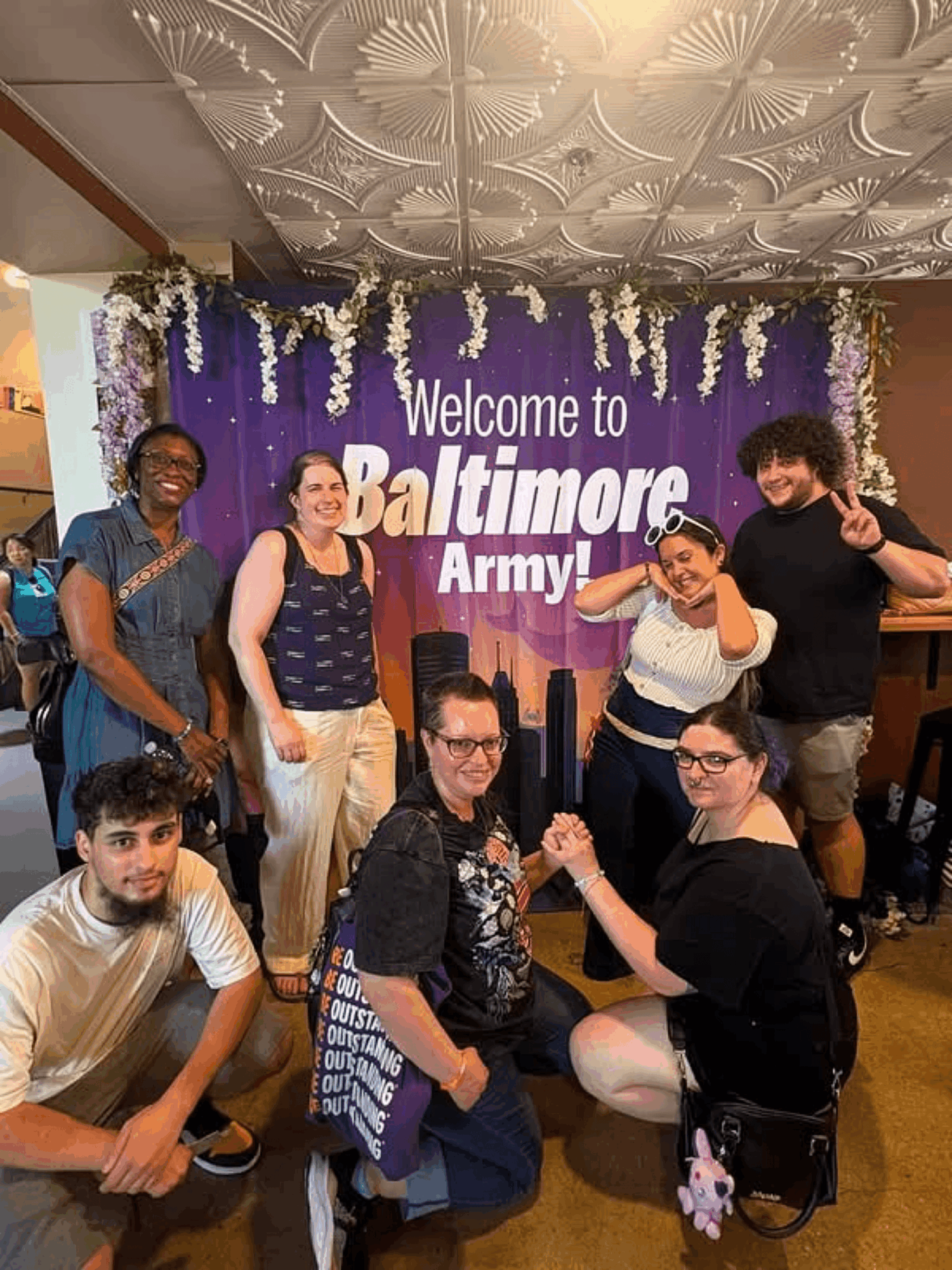 Group of seven people posing in front of a purple Baltimore Army welcome banner, some making fists, indoors with decorative ceiling.