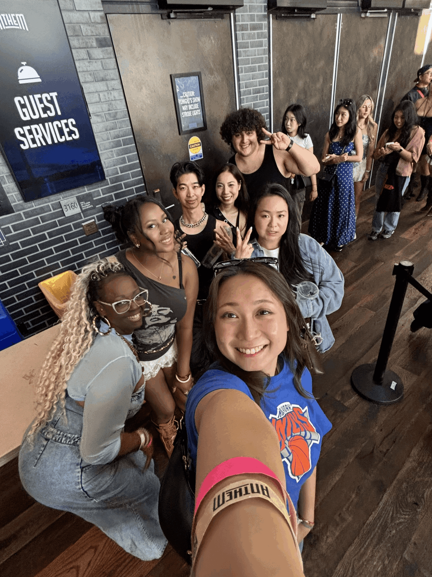 Group of people smiling and taking a selfie indoors, with a line and guest services sign in the background.